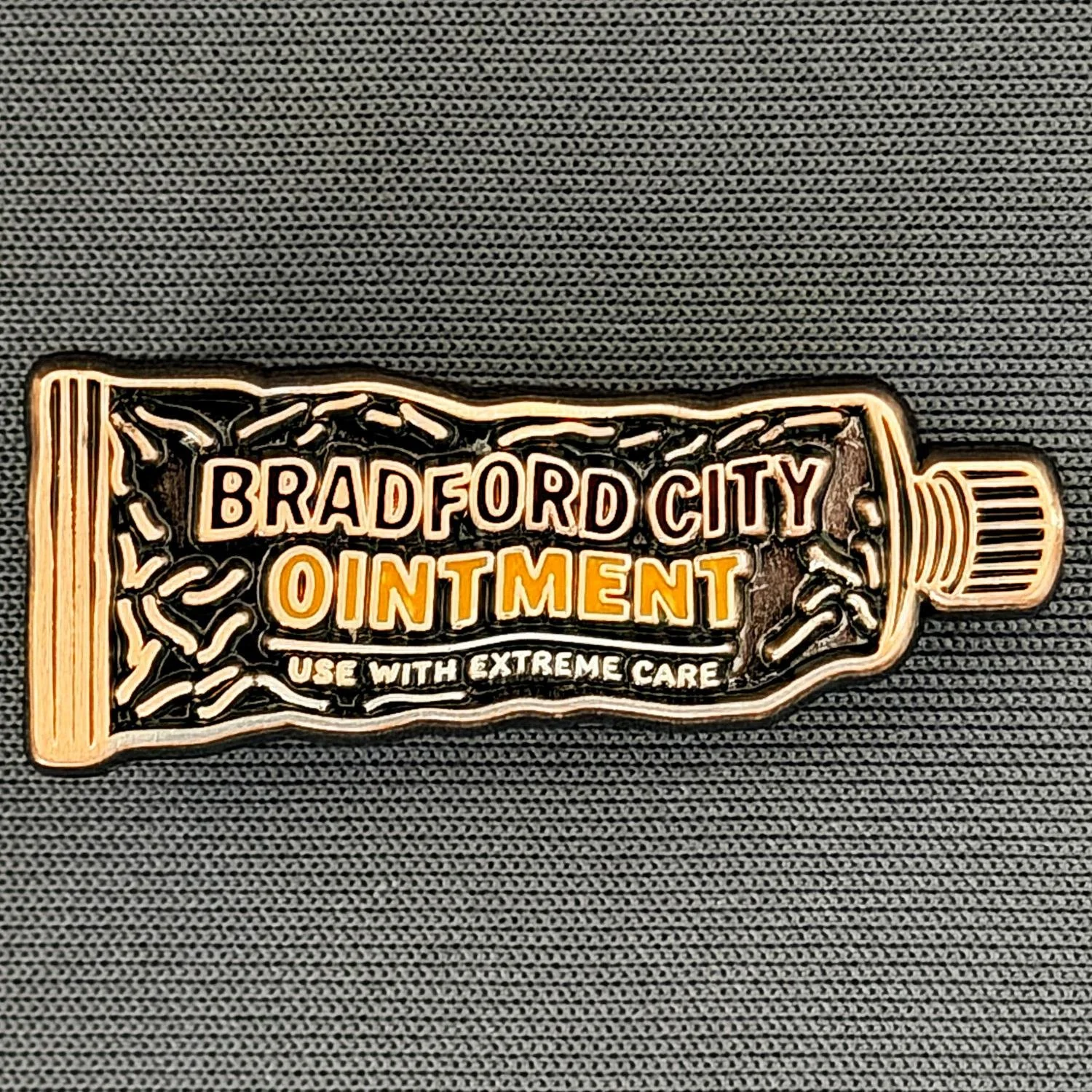 A patch designed to look like a tube of ointment with the text "Bradford City Ointment" and "Use with Extreme Care" on it, placed on a textured black fabric background.