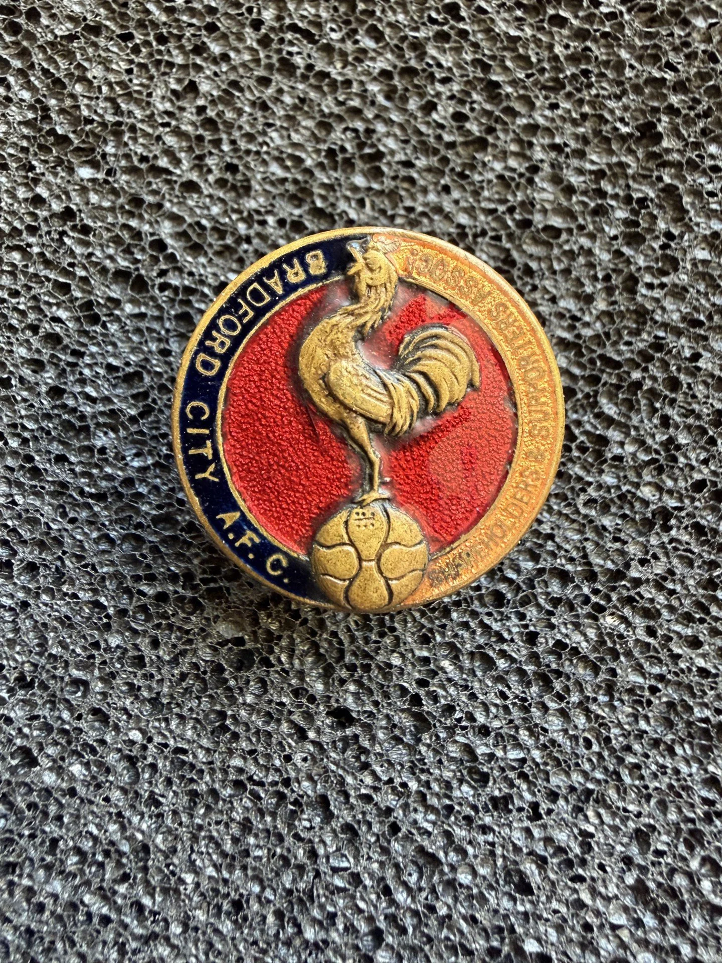 Close-up of a circular metal badge with a rooster in the center, surrounded by blue, red, and gold colors, with text around the edge on a textured dark background.