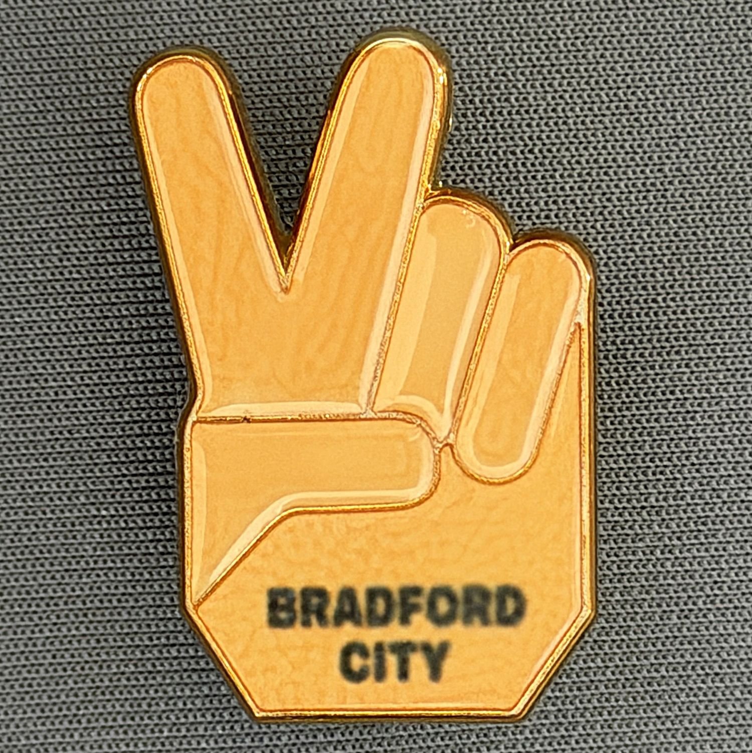 Gold-colored lapel pin in the shape of a hand making a peace sign, with the words "Bradford City" on it, pinned on a textured gray fabric.