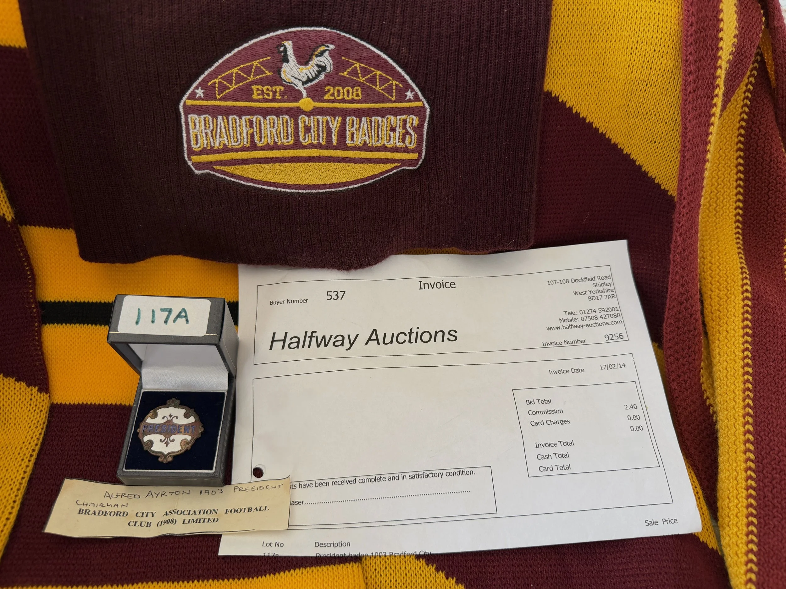 Close-up of a Bradford City Badge, a football club badge, on a maroon and yellow striped sports jersey. Also visible is an auction invoice, a pin with a presidential emblem, and a handwritten note.