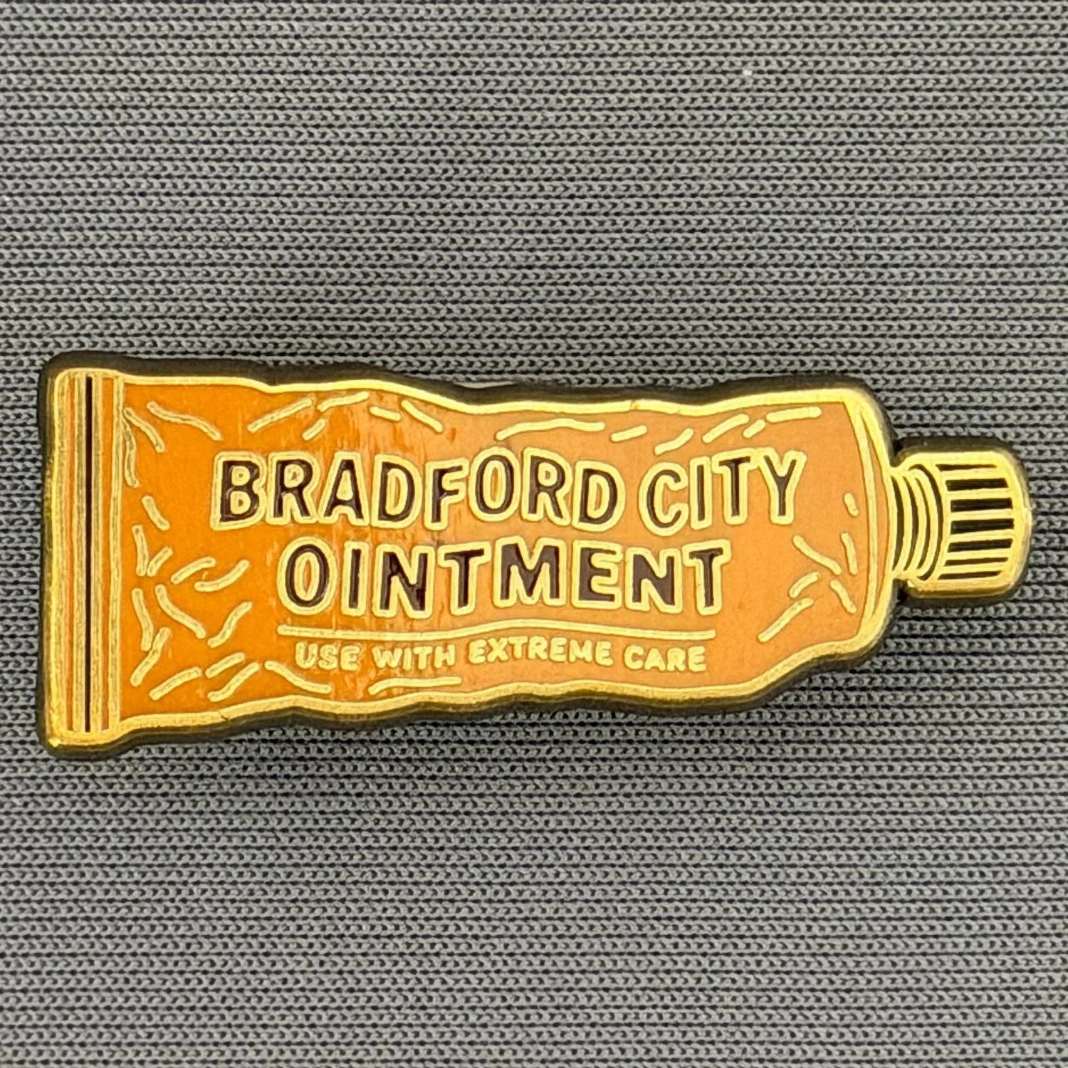 A pin shaped like a tube of ointment with the text 'Bradford City Ointment, Use with Extreme Care' on it, placed on a textured gray surface.