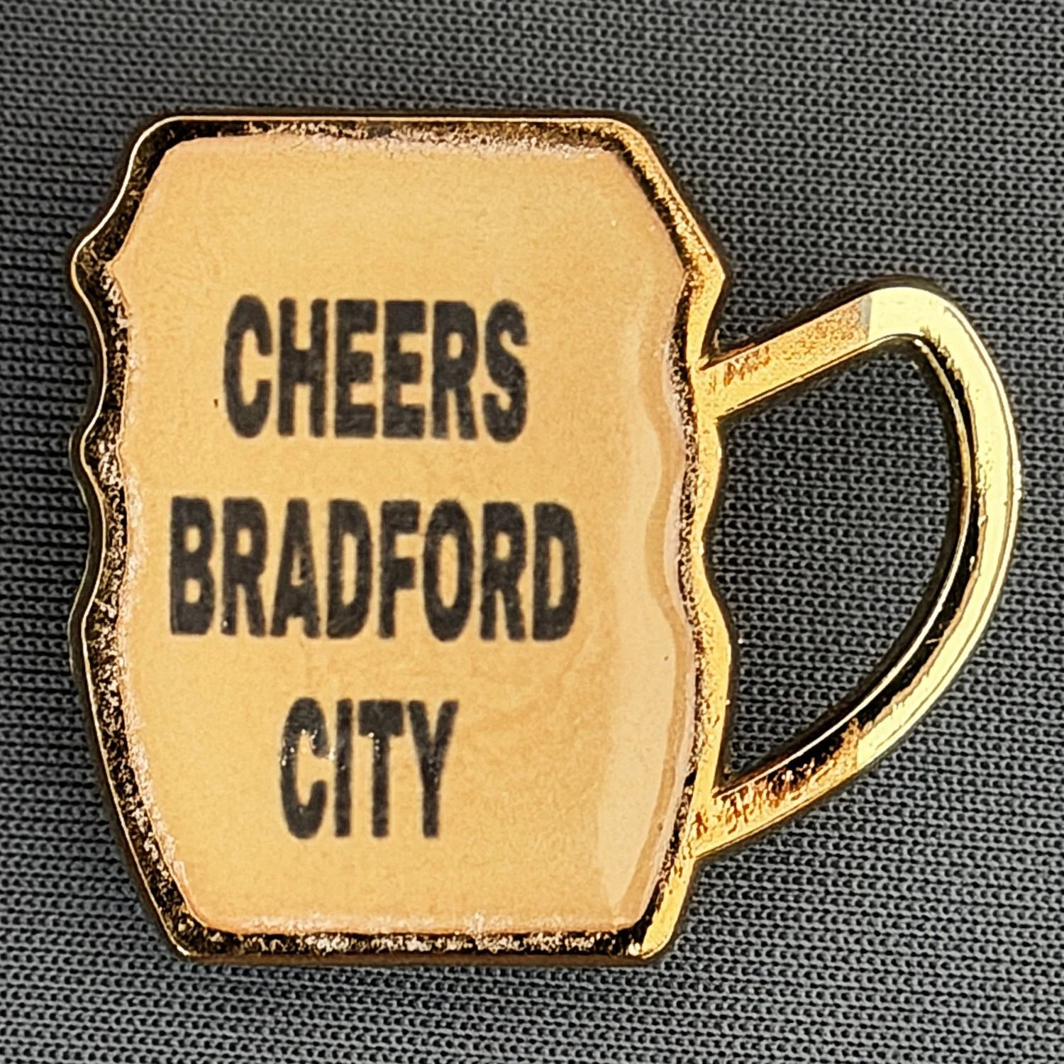 A gold-colored keychain in the shape of a coffee mug with the words 'Cheers Bradford City' written on it, placed on a dark fabric background.