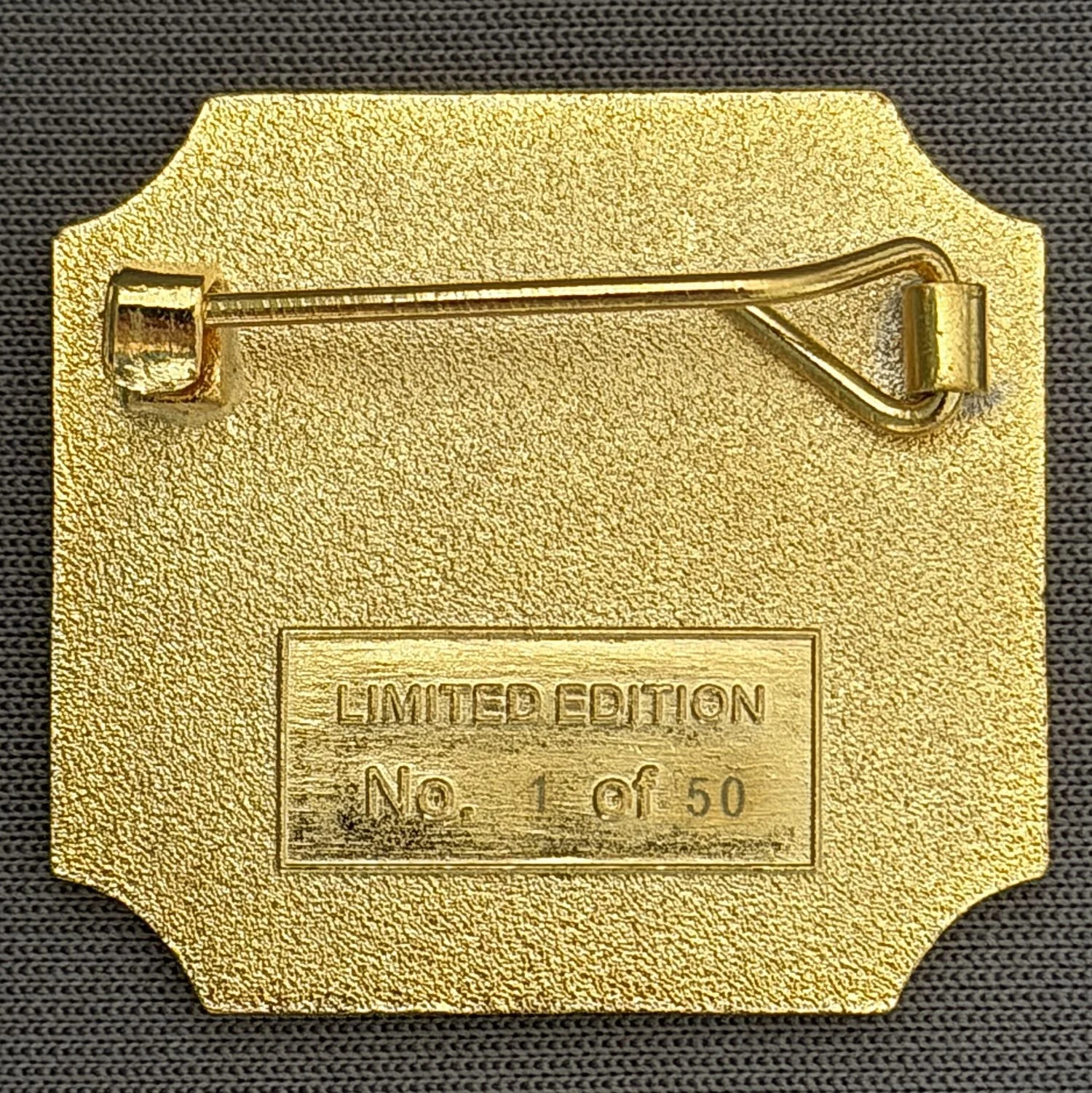 Gold-colored limited edition badge or plaque with a pin on the back, resting on a dark textured surface.