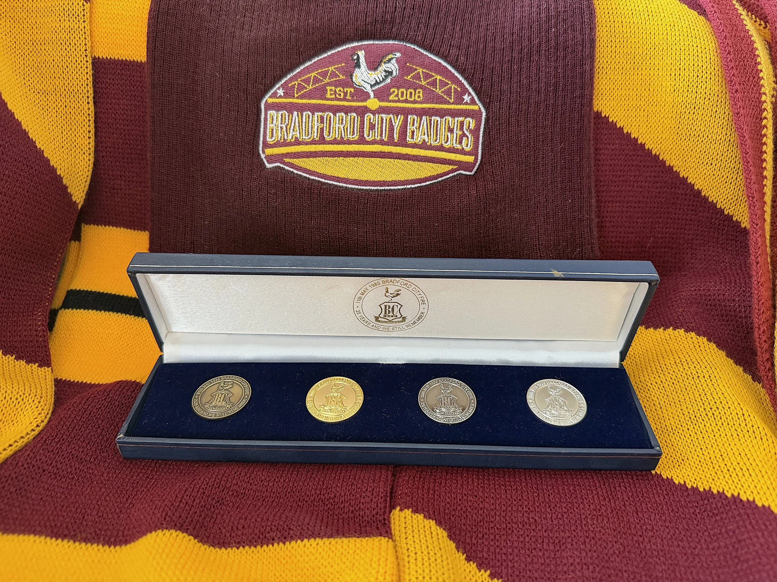 A maroon and gold sports jersey with a patch reading 'Bradford City Badgers EST 2008' and a crest with a rooster, with a display case underneath containing five coins with emblems related to Bradford City.