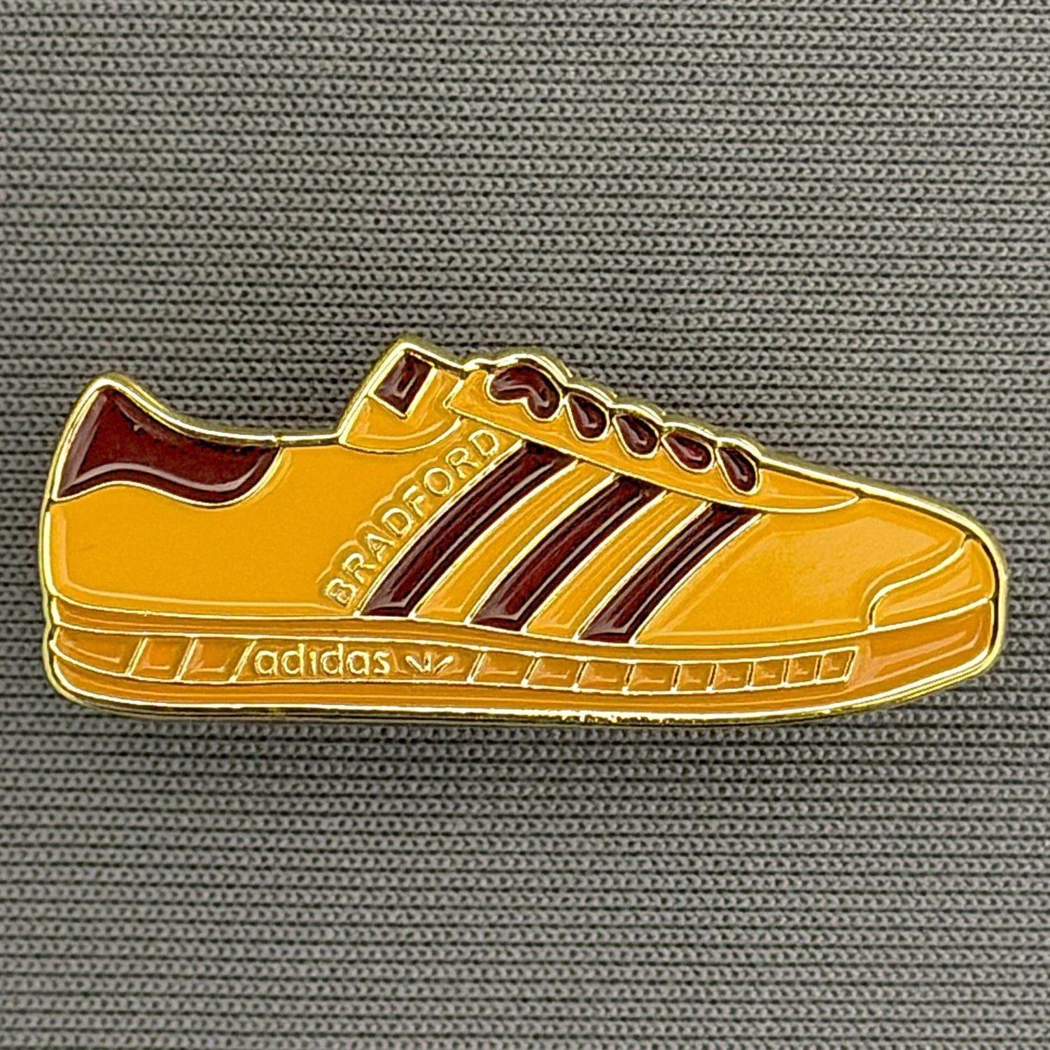 Reference Number: BCUN044
Size: H 12.2 mm x W 30.4 mm
Pin Type: Brooch Pin
Plating: Gold
Value: £5
Manufacturer: Unknown
Comments: Amber & Claret Trainer, Large Version