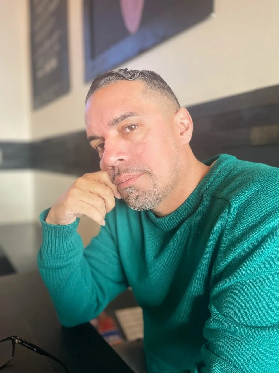 A man with short dark hair and a beard, wearing a teal sweater, sitting at a table in a cafe or restaurant, resting his chin on his hand, looking at the camera.