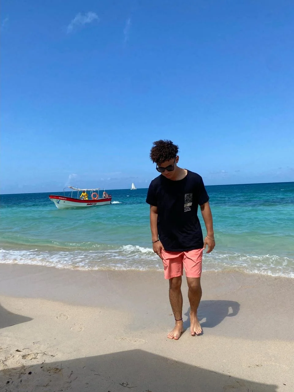 A young man in black t-shirt and pink shorts walking barefoot on a sandy beach near the ocean with a boat and sailboat in the background under a blue sky.