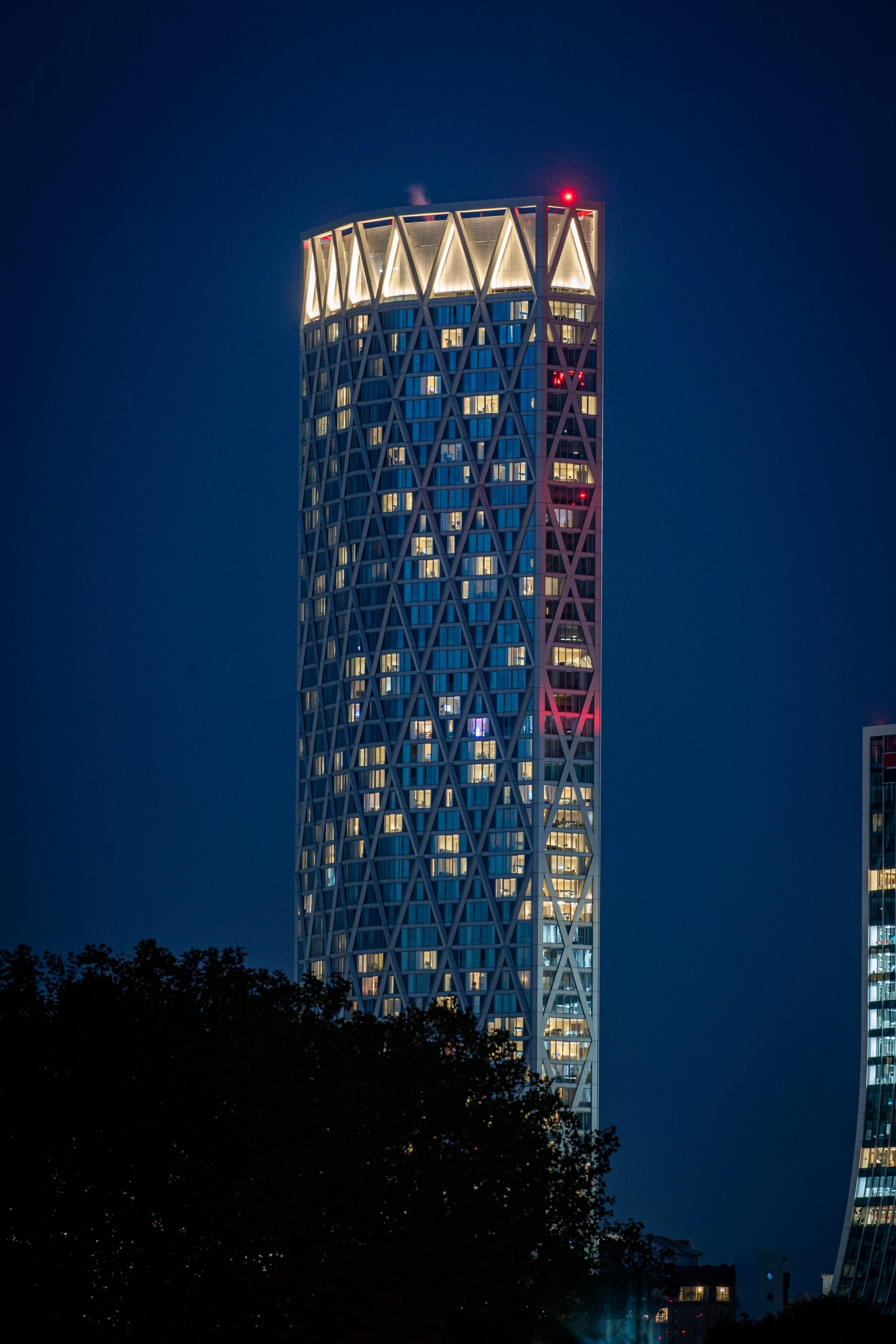 Nighttime image of a skyscraper with a unique lattice exterior and illuminated windows, with a clear sky and some trees at the bottom.