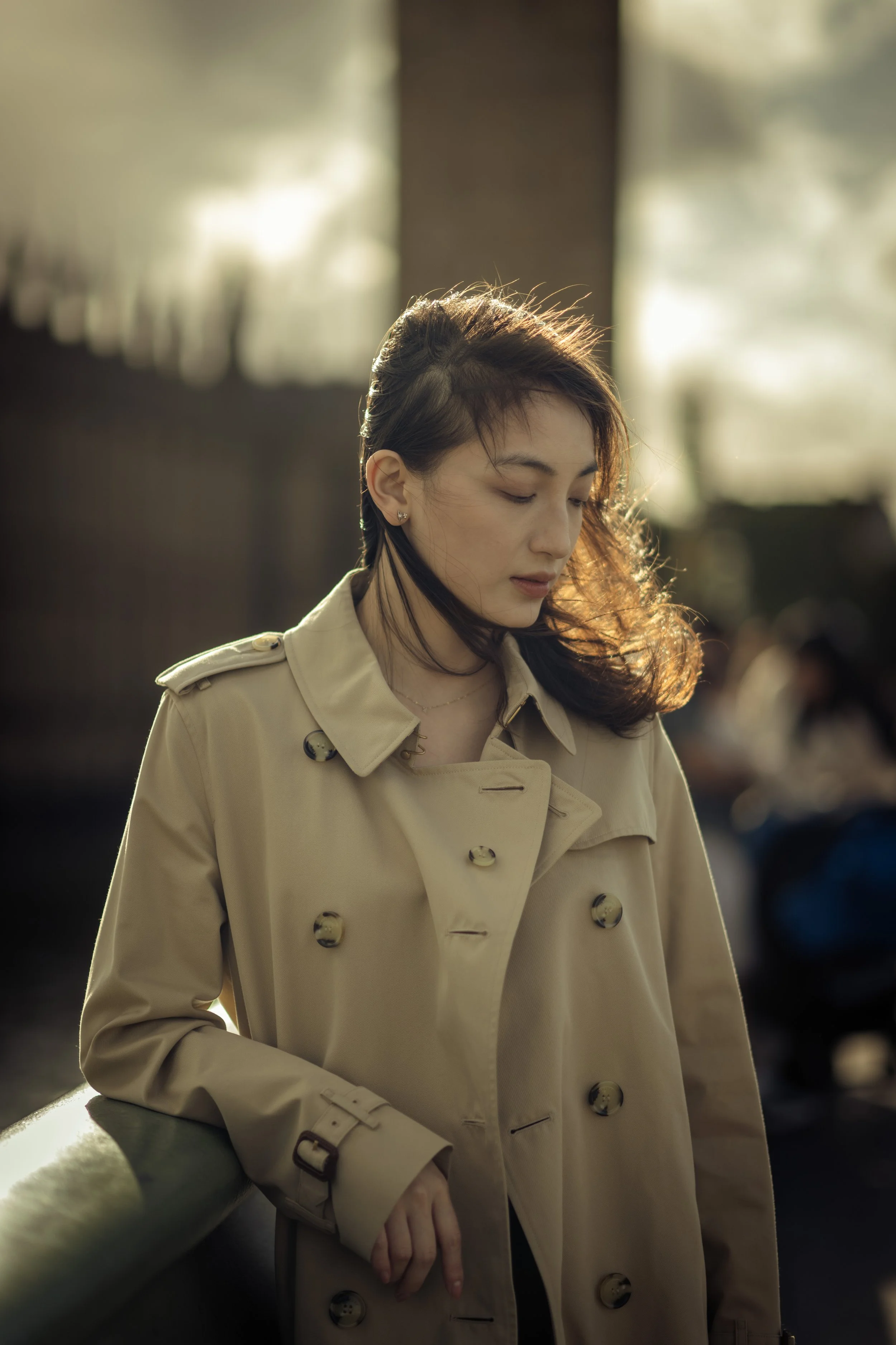 A young woman in a beige trench coat standing indoors, with sunlight illuminating her from behind, her eyes closed and head slightly bowed.