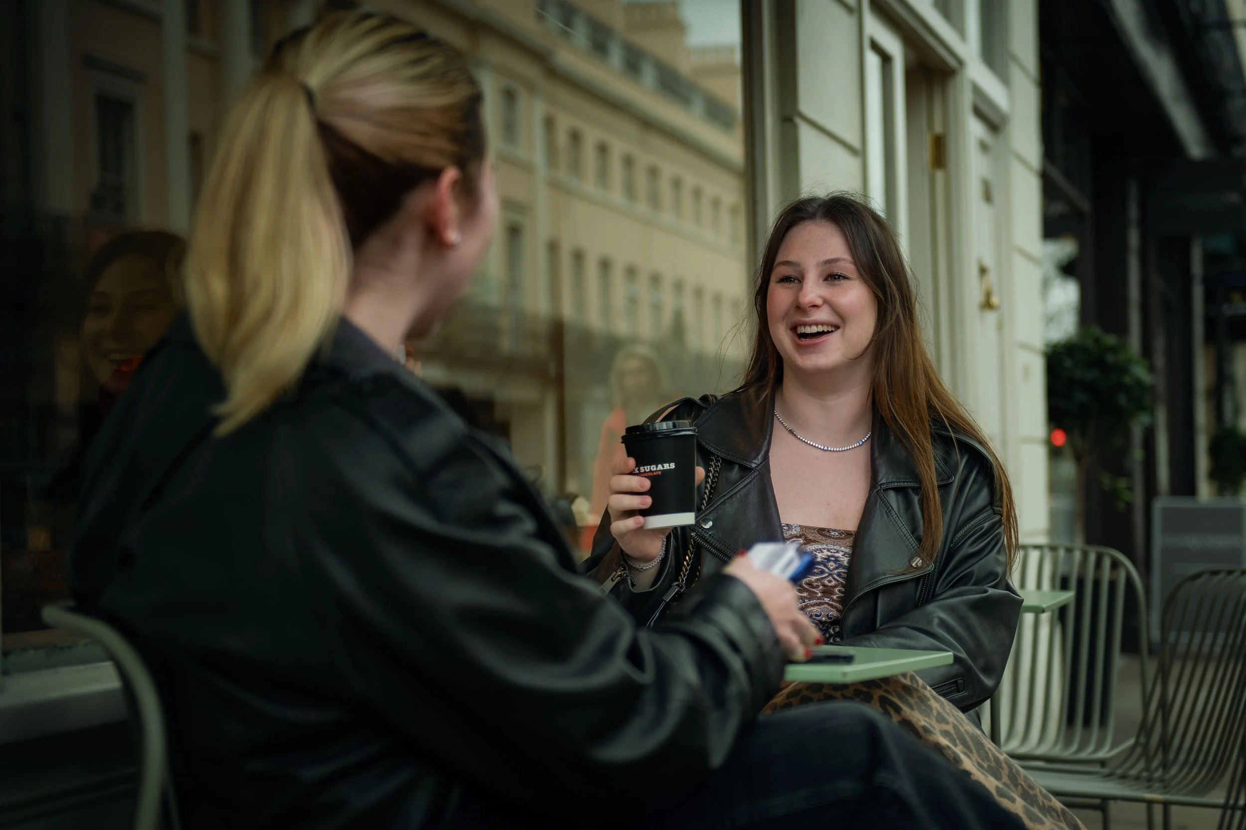 Two women sitting outside a cafe talking, one holding a cup of coffee, the other holding a phone.