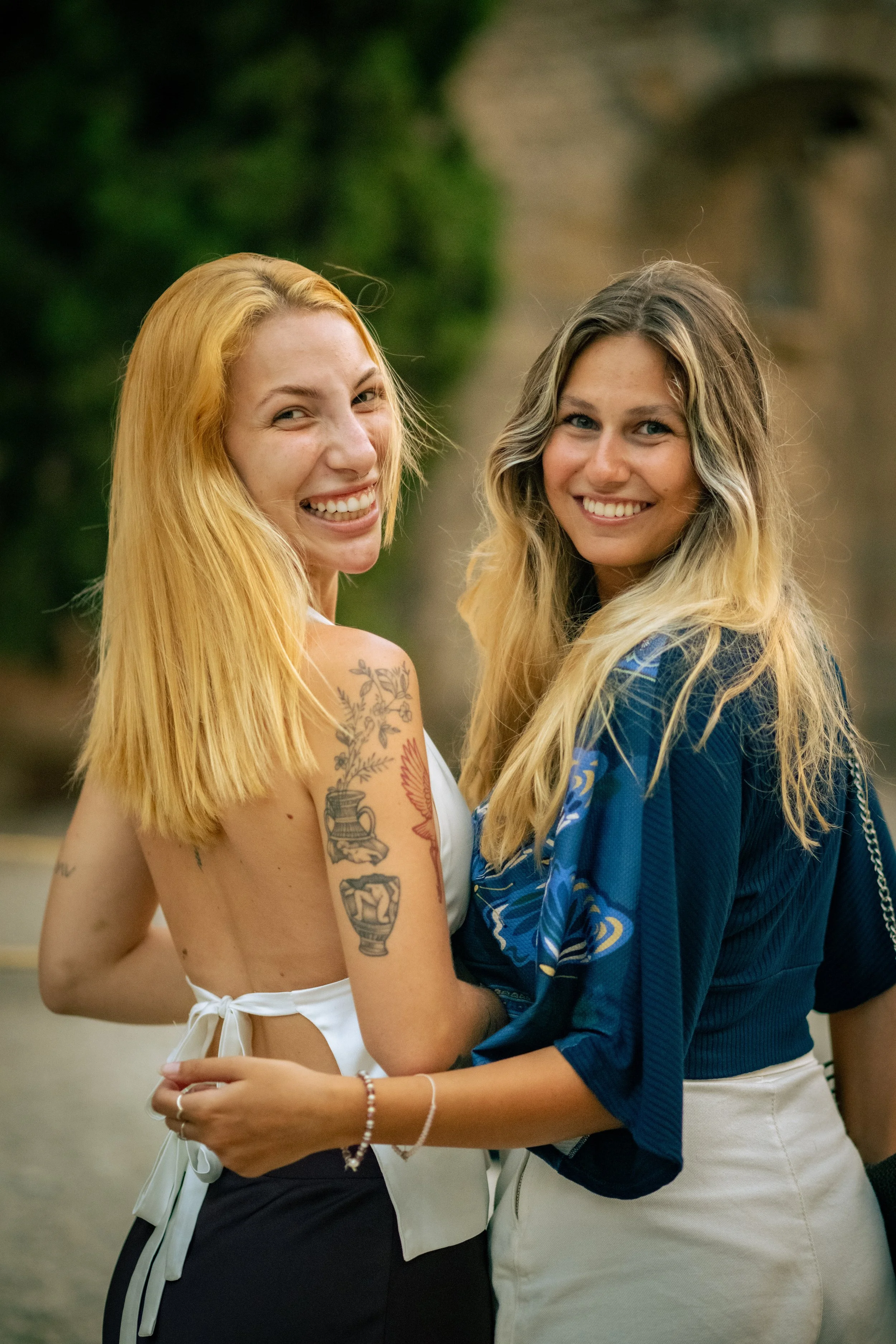 Two young women hugging and smiling outdoors, with one showing tattoos on her shoulder.
