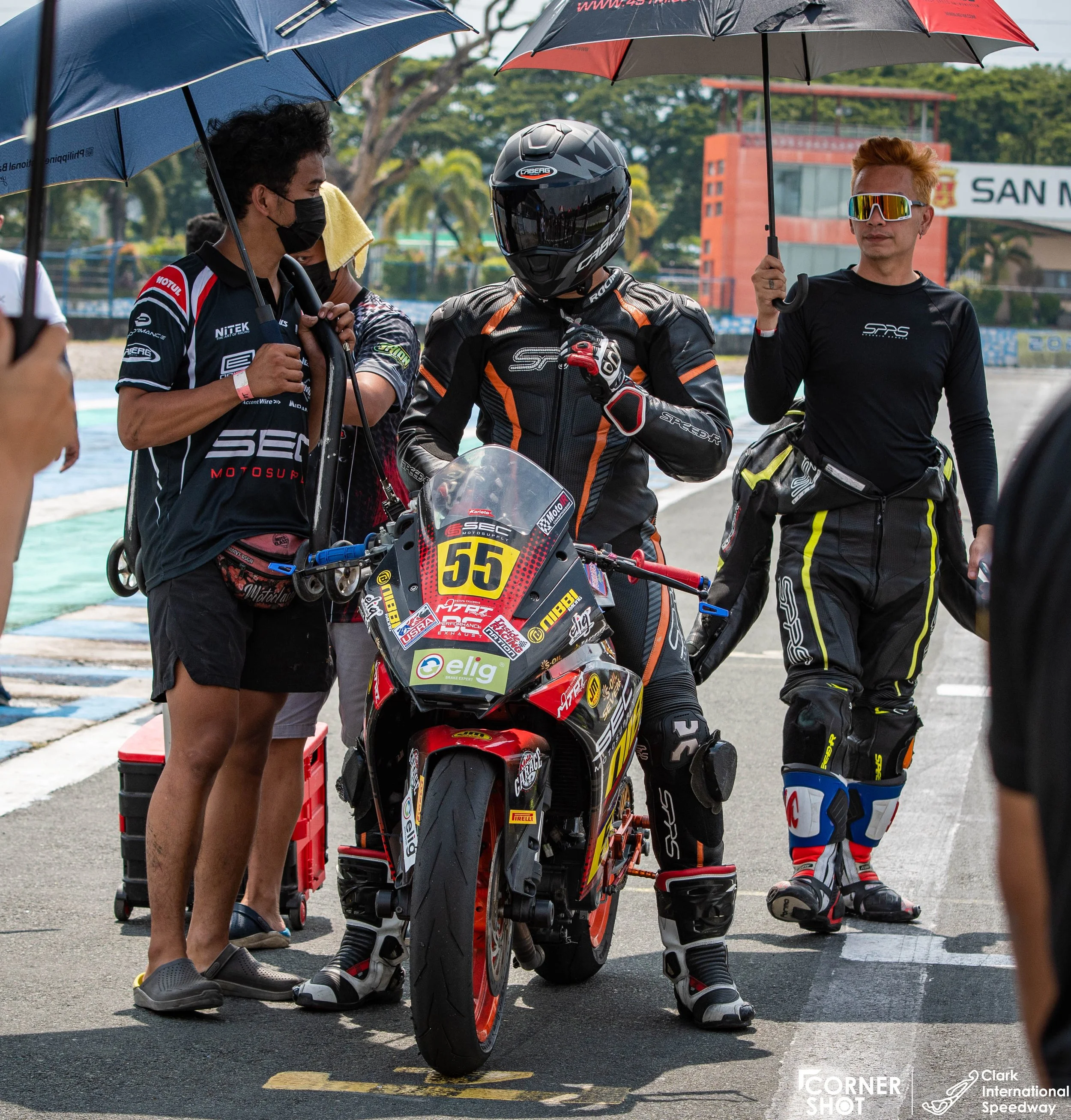 Lawrence Valencia in full gear standing beside a race bike with the number 55 on it, team members holding umbrellas over him at a racetrack. Other team personnel in racing suits and face masks are also present.