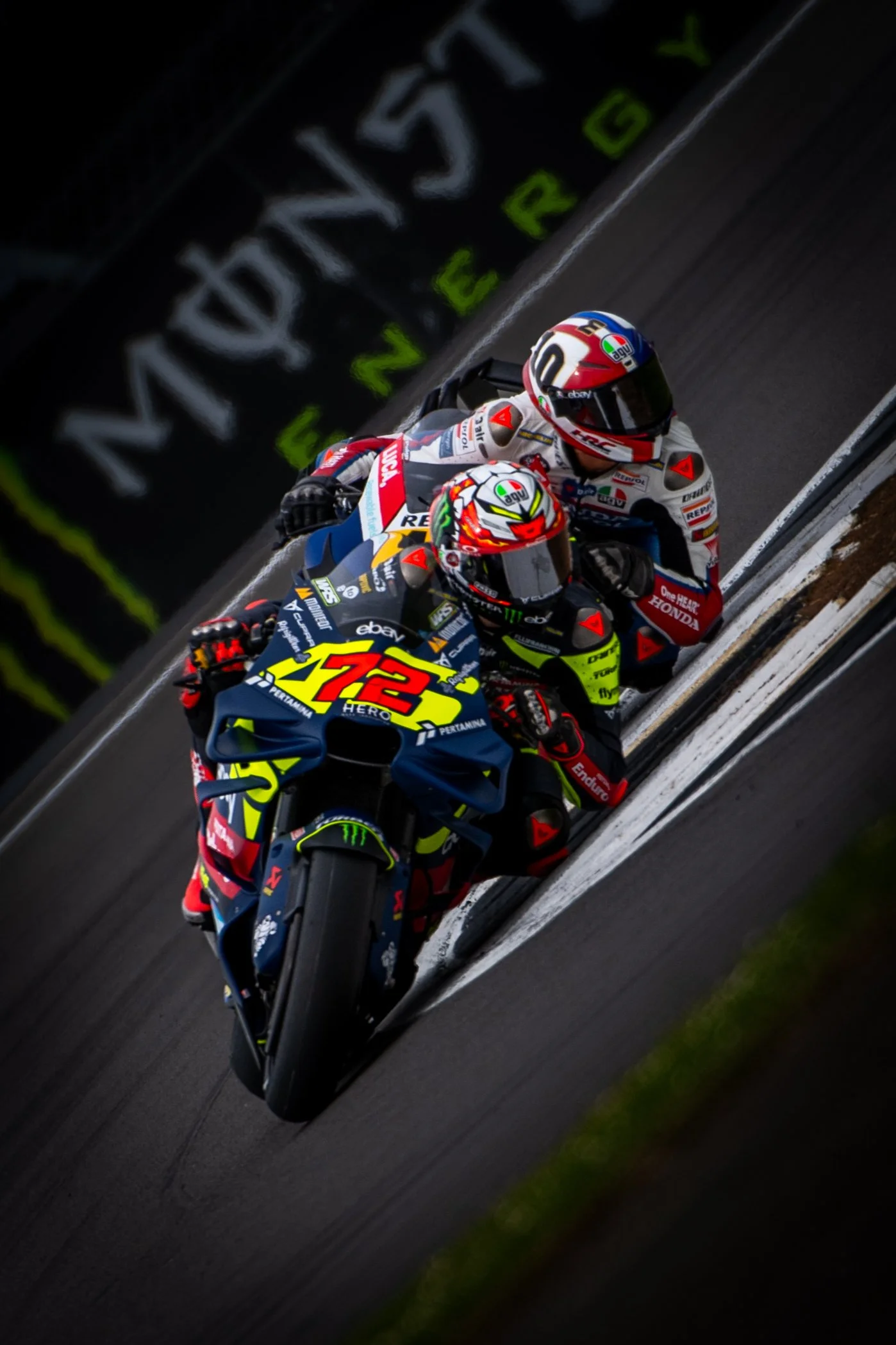 Two motorcycle racers in racing suits and helmets riding on a racetrack, with one rider leading and the other closely following.