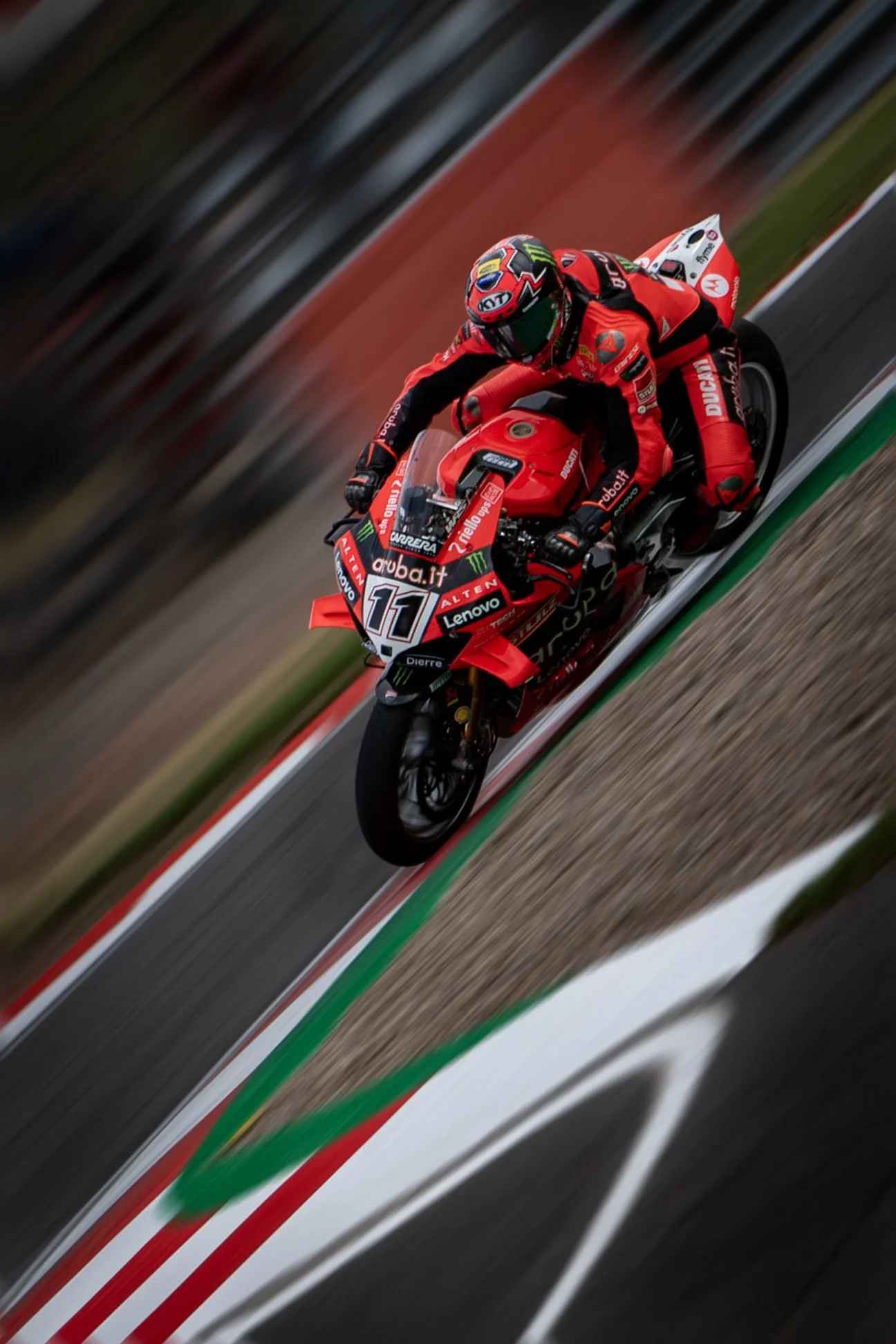 A professional motorcycle racer in red racing gear leaning into a turn on a race track, riding a red Ducati motorcycle with sponsor logos, blurred background indicating high speed.