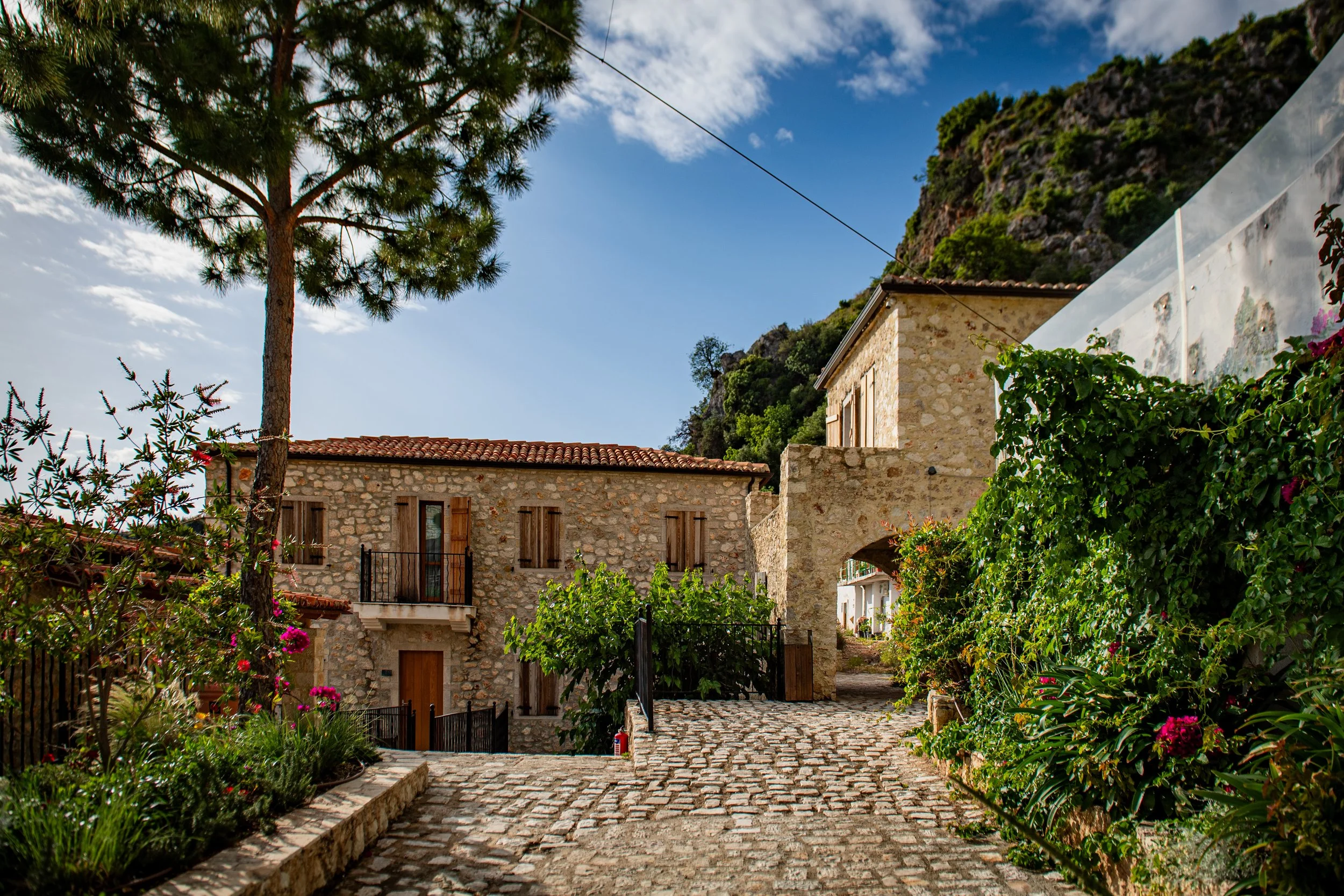 Stone houses with red tile roofs on cobblestone path surrounded by greenery, trees, and flowering bushes under a partly cloudy sky.