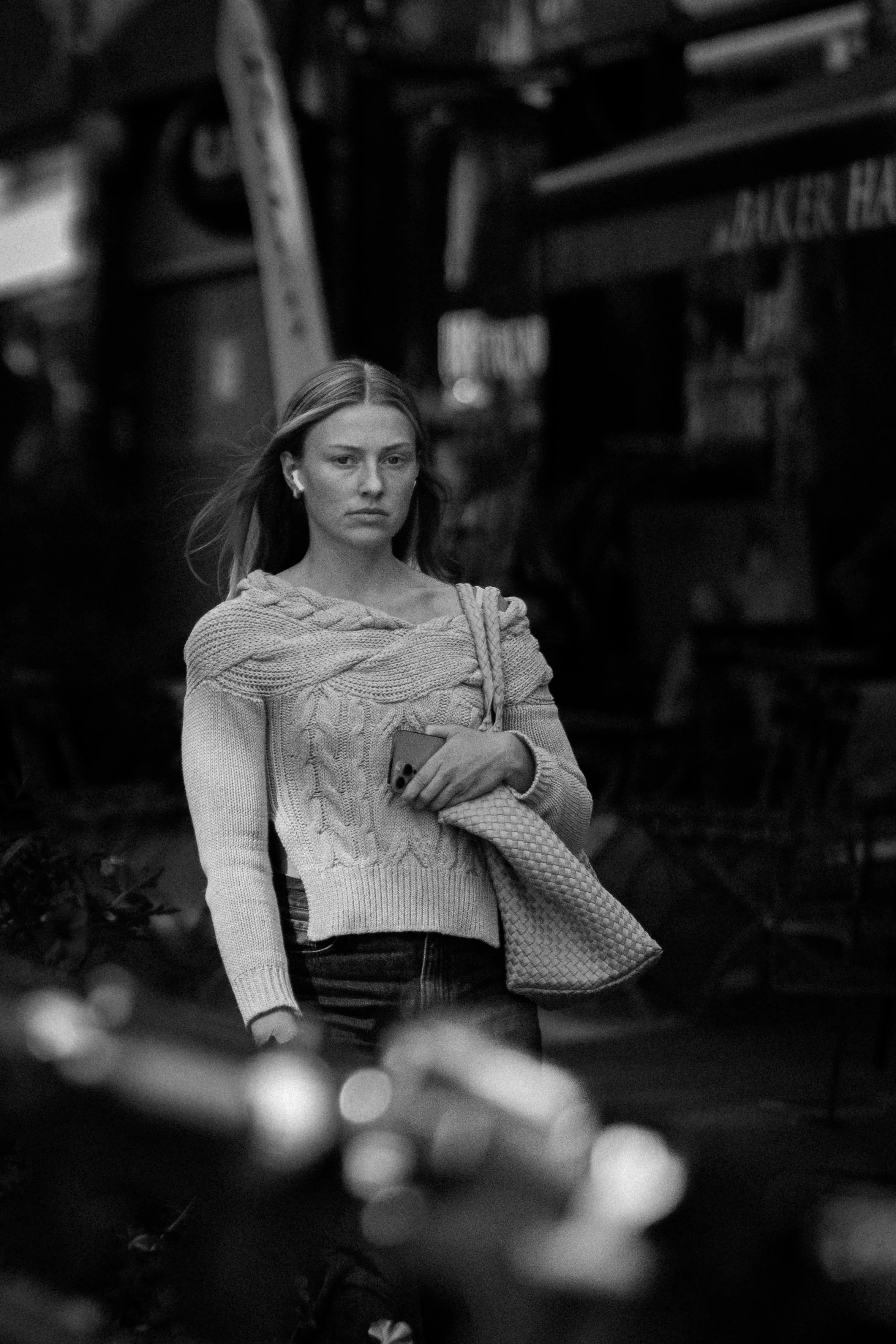 A woman with long hair, wearing a knitted sweater, standing outdoors at night with a serious expression, holding a phone and a woven bag.