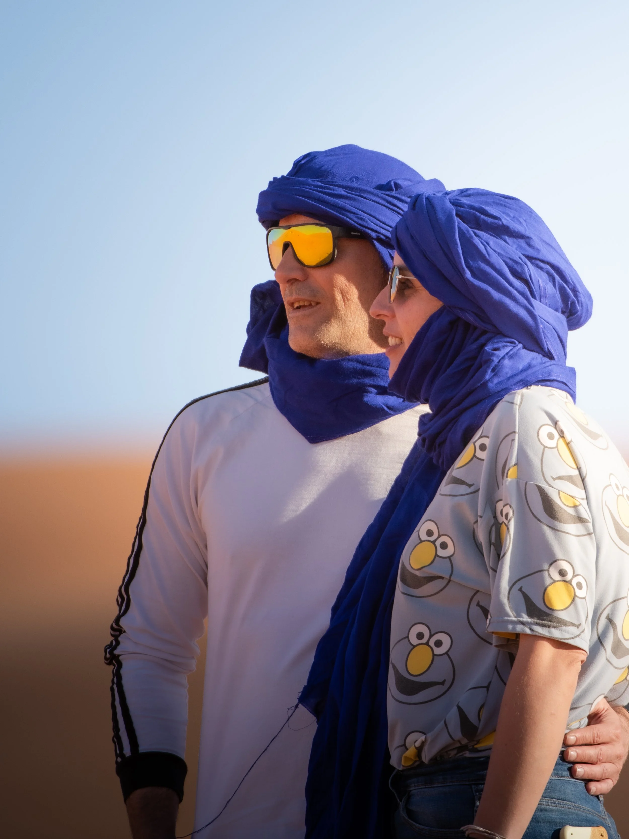 A man and woman wearing blue headscarves and sunglasses, standing outdoors in a sandy or desert environment, smiling and looking into the distance.