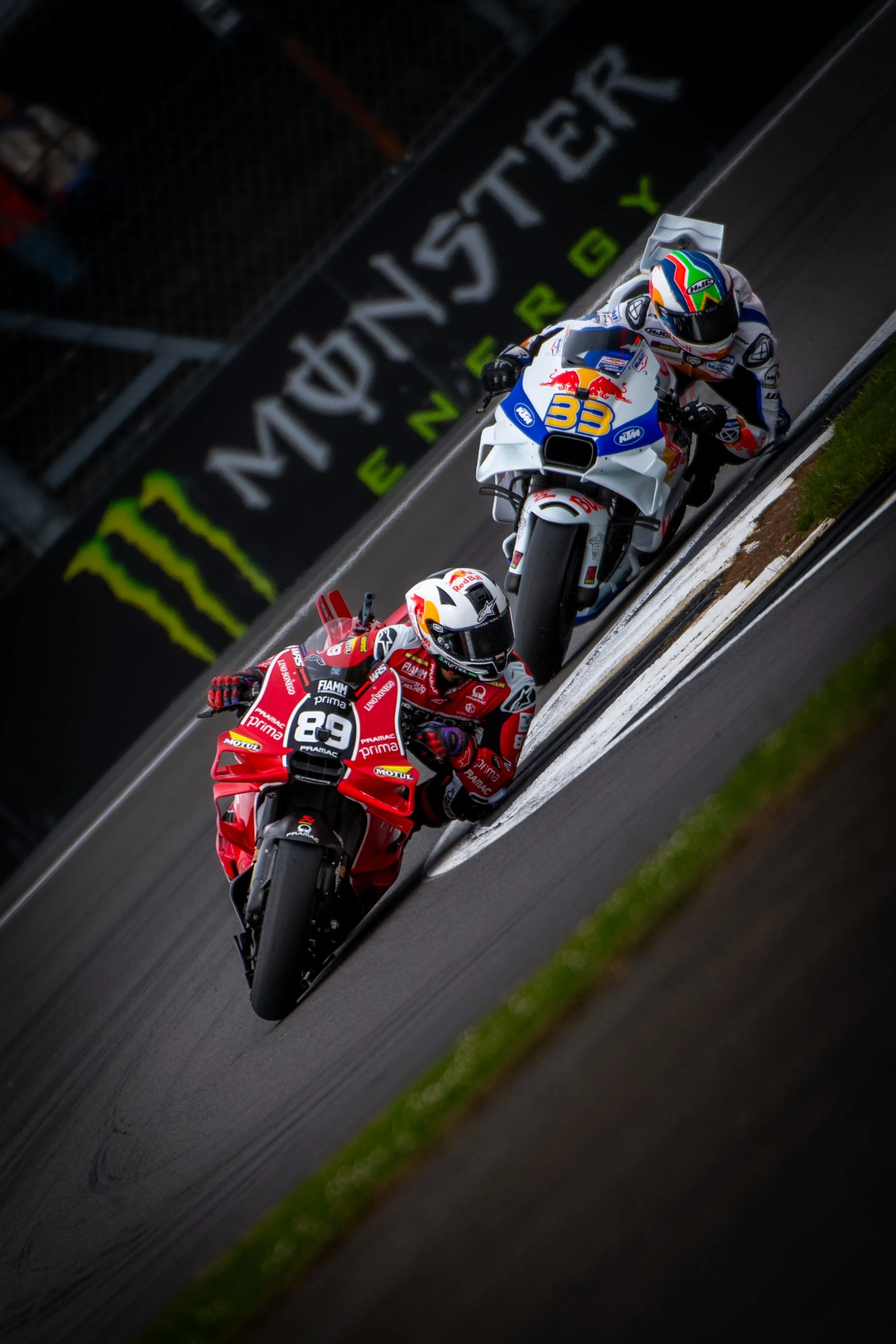 Two motorcycle racers leaning into a turn on a racetrack, one in red and the other in white and blue, with Monster Energy branding in the background.