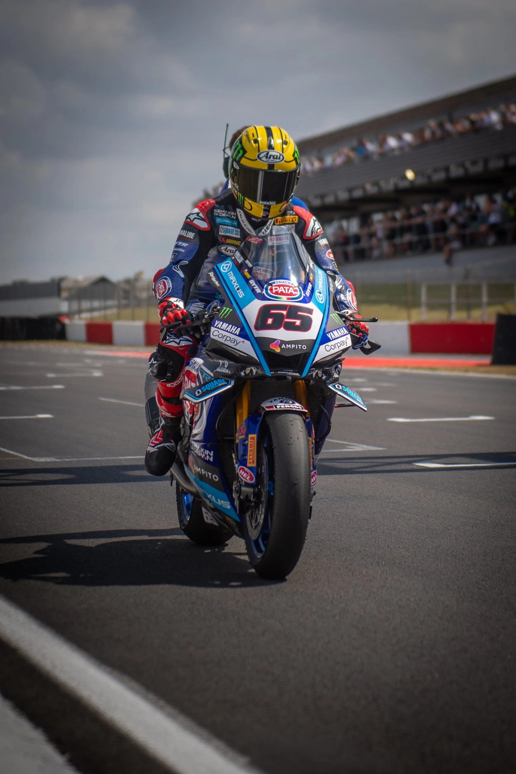 A motorcycle racer in a racing suit with a yellow helmet riding on a race track with grandstands and spectators in the background.