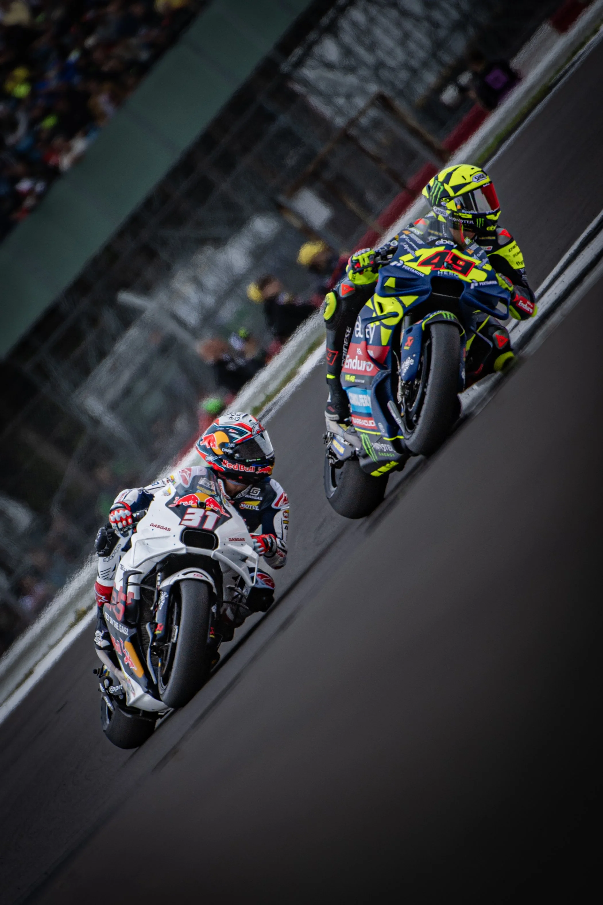 Two professional motorcycle racers in racing suits and helmets riding on a racetrack during a competition, with spectators and railings in the background.