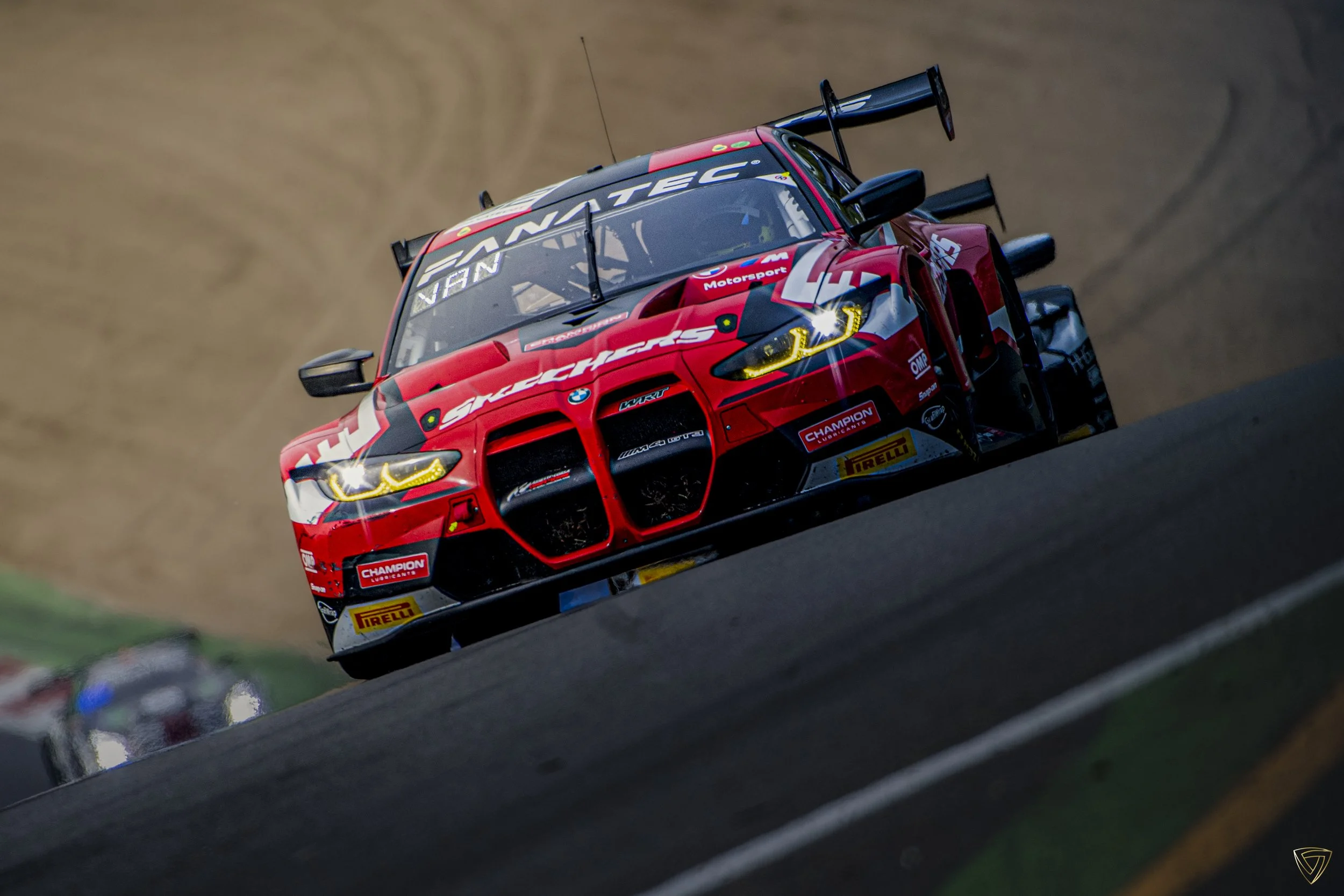 A red race car with BMW and Pirelli branding, racing on a track, with a blurred background and another car behind it.