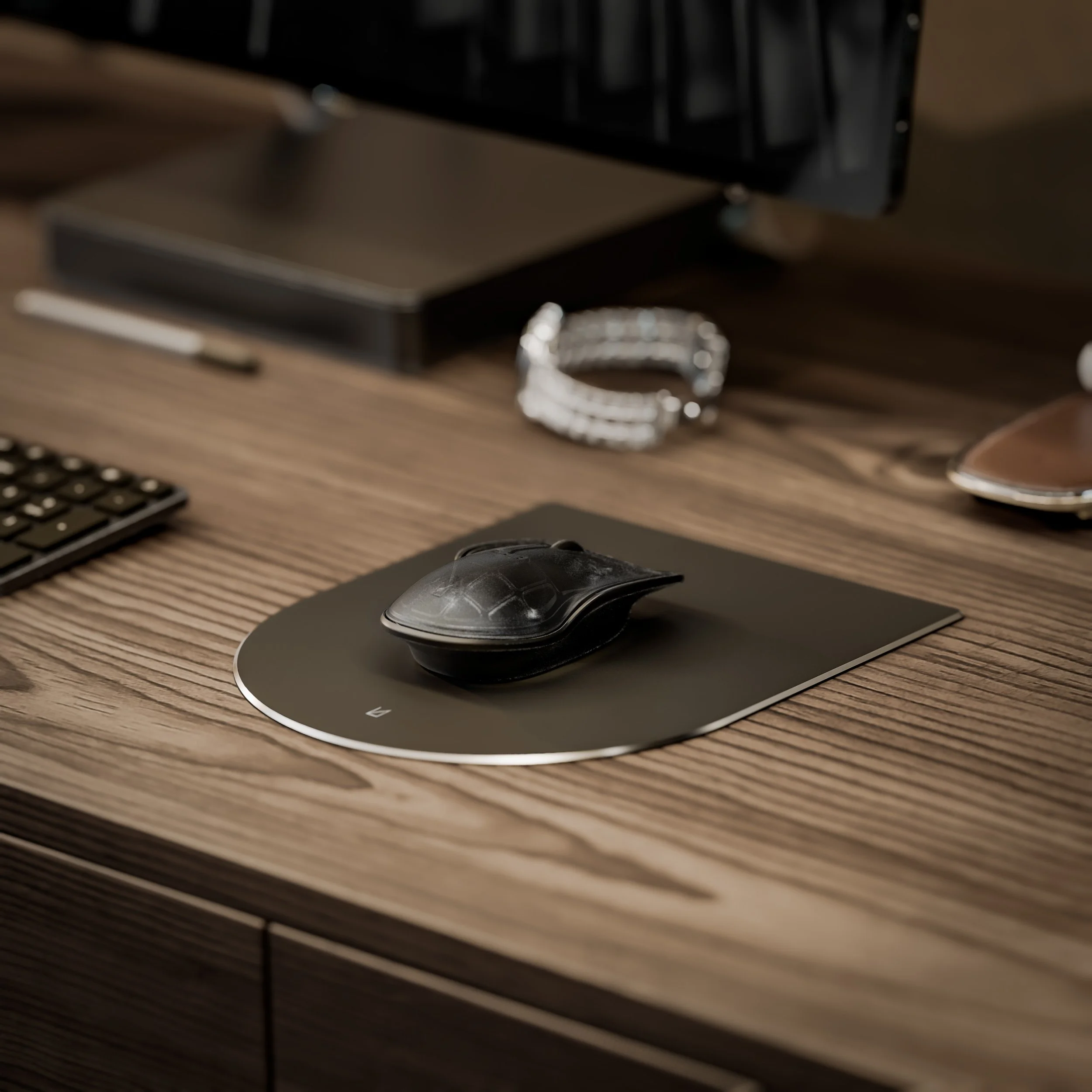 Close-up of a computer mouse on a black mouse pad on a wooden desk, with a keyboard, pen, wristwatch, and other office items in the background.