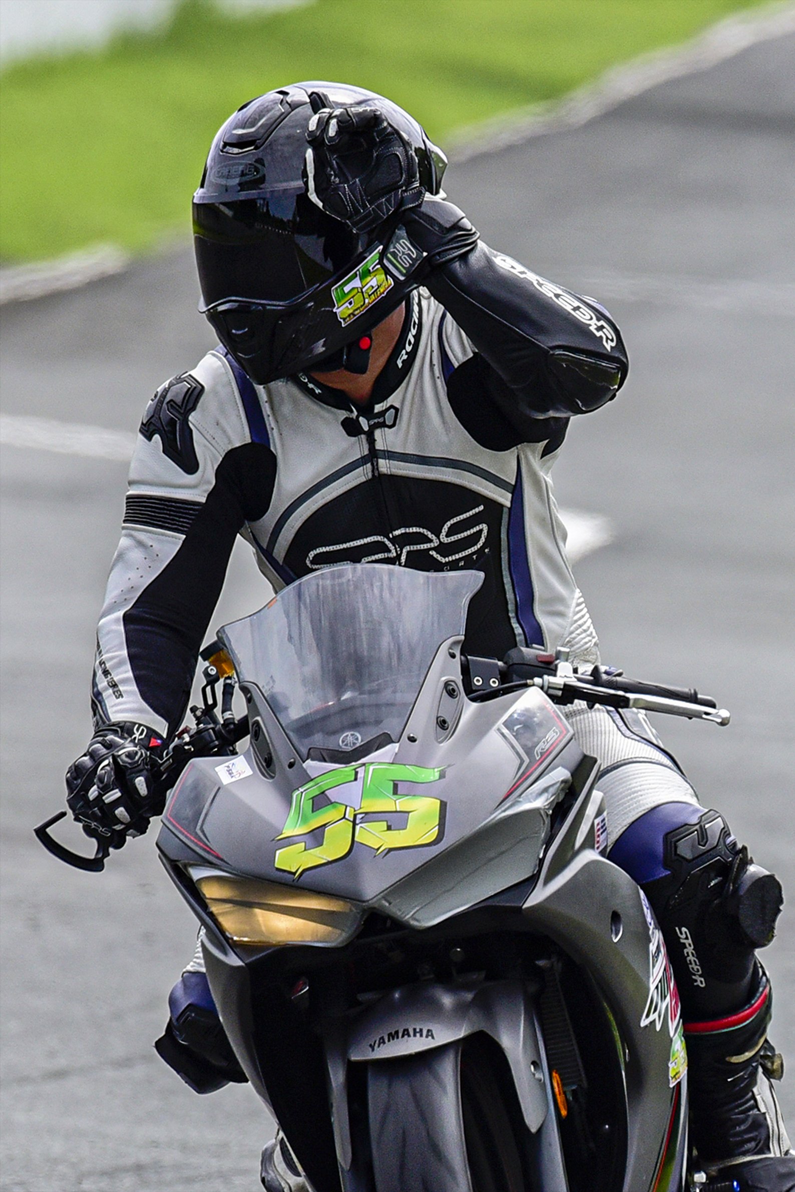 A motorcycle racer in full gear adjusts their helmet while sitting on a Yamaha racing bike with the number 55 on the front, on a racetrack. rider Lawrence Valencia