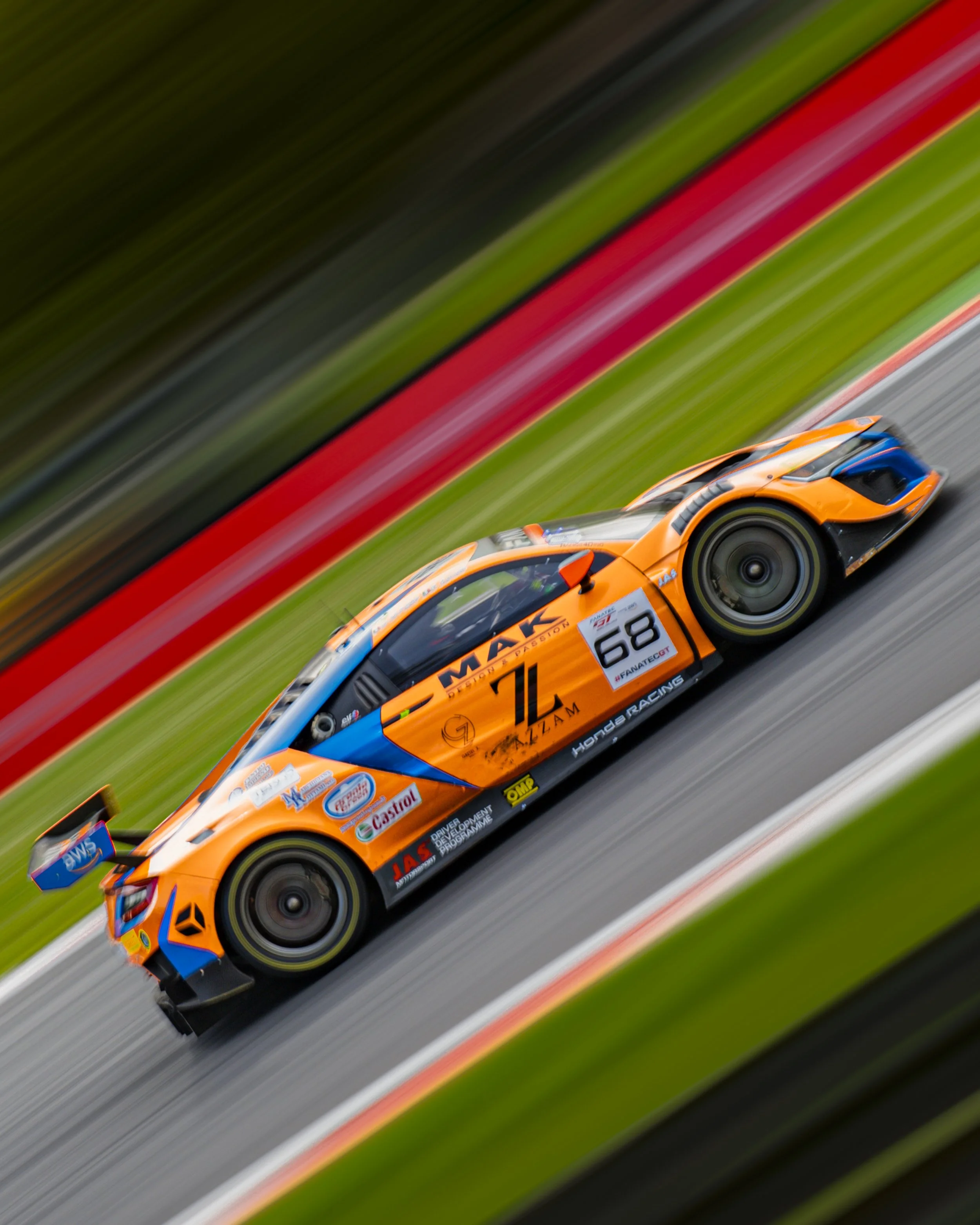 An orange racing car with the number 68 on the side speeding on a racetrack with blurred background.