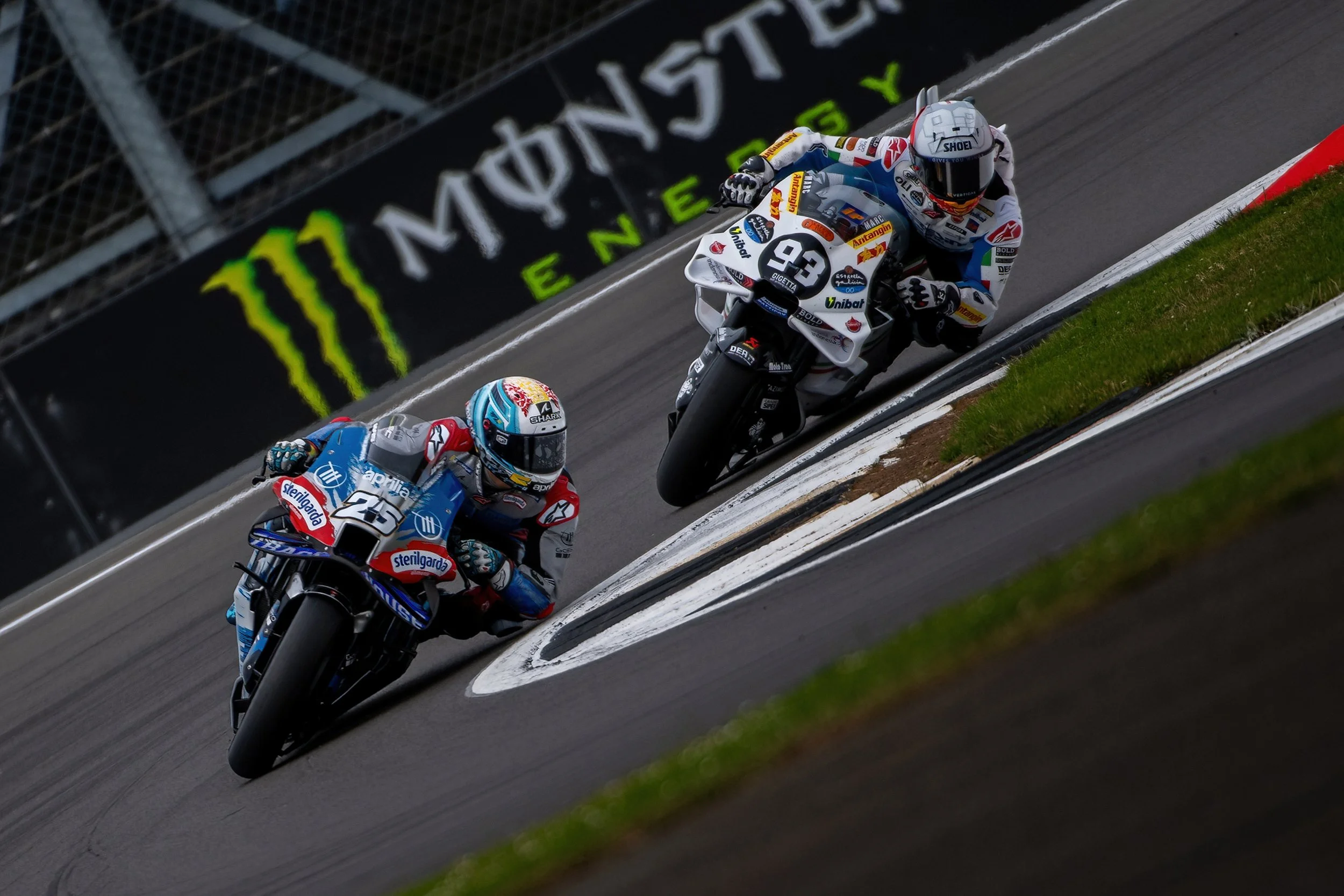Two motorcycle racers leaning into a curve on a racetrack, with sponsor banners and greenery along the edge.