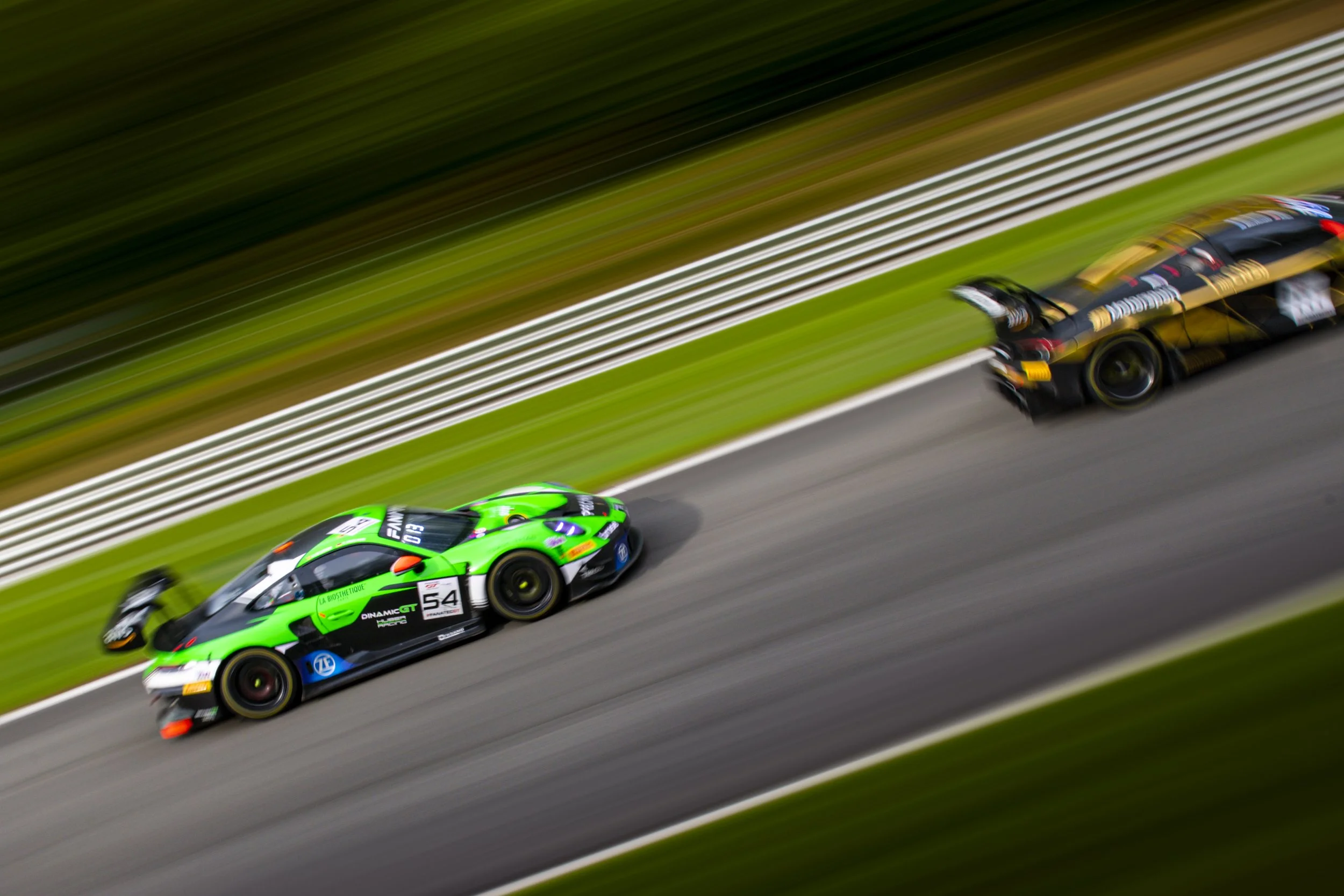 Race cars speeding on a racetrack with blurred background, one green and black car in the foreground and a black and gold car behind.