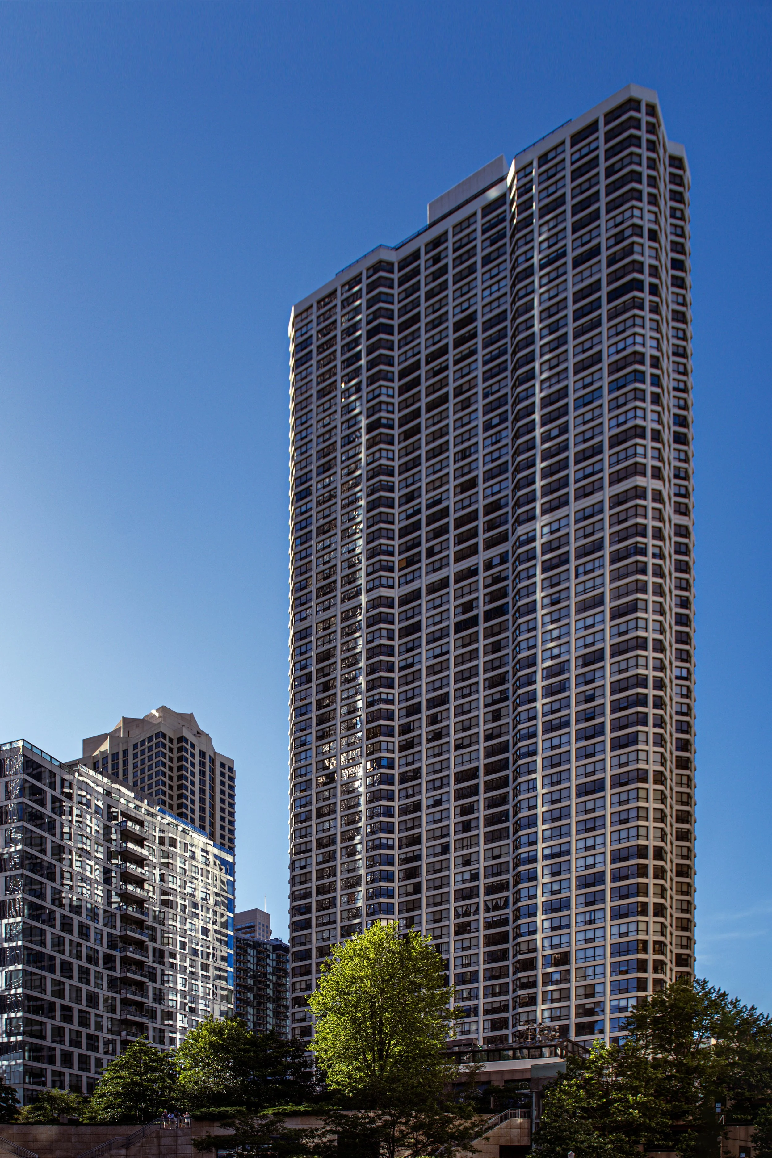 Tall modern skyscraper in a city, with a blue sky background and green trees at the base.