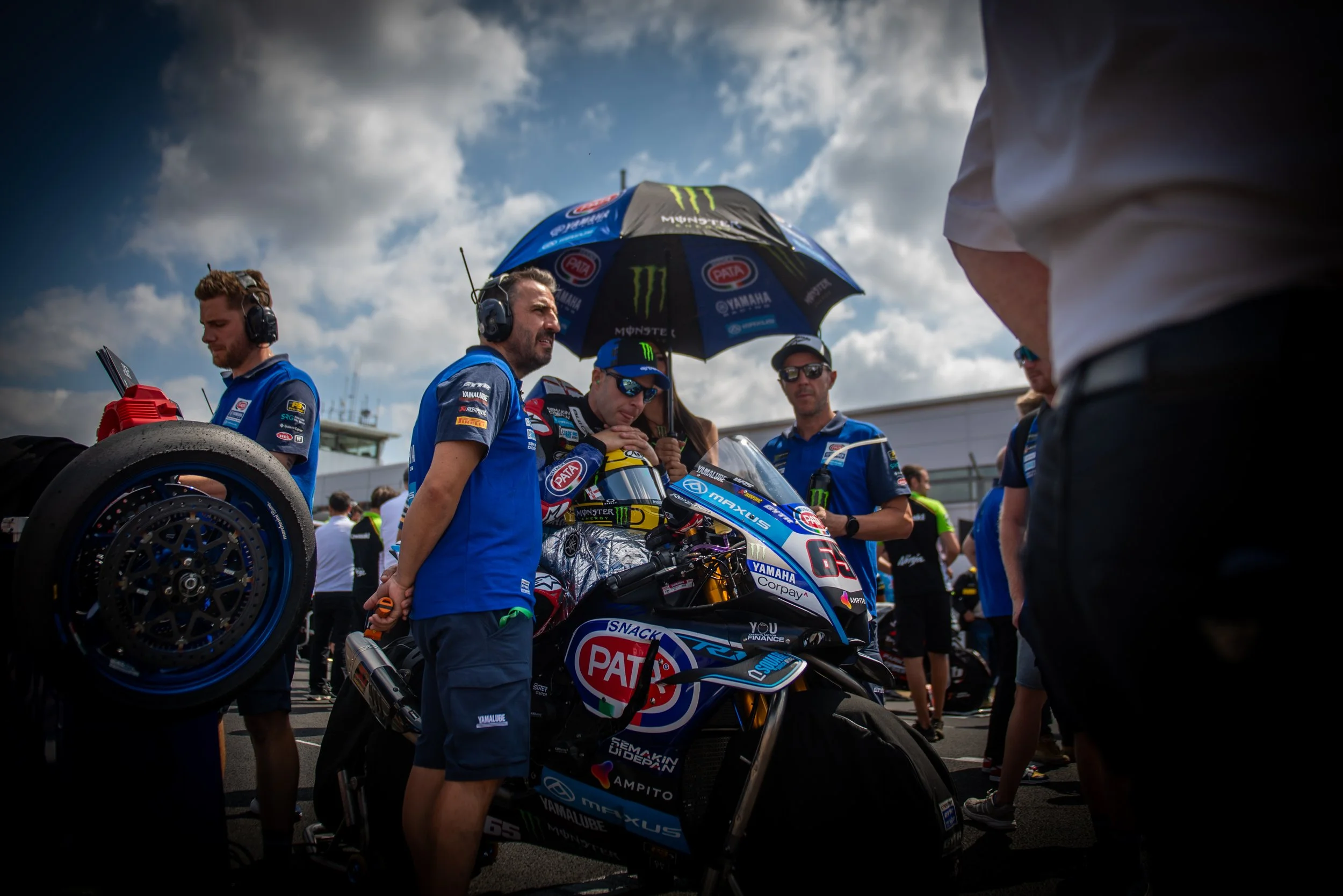 Motorcycle racer preparing in a track pit area with team members around, holding an umbrella over the rider, with cloudy sky above.