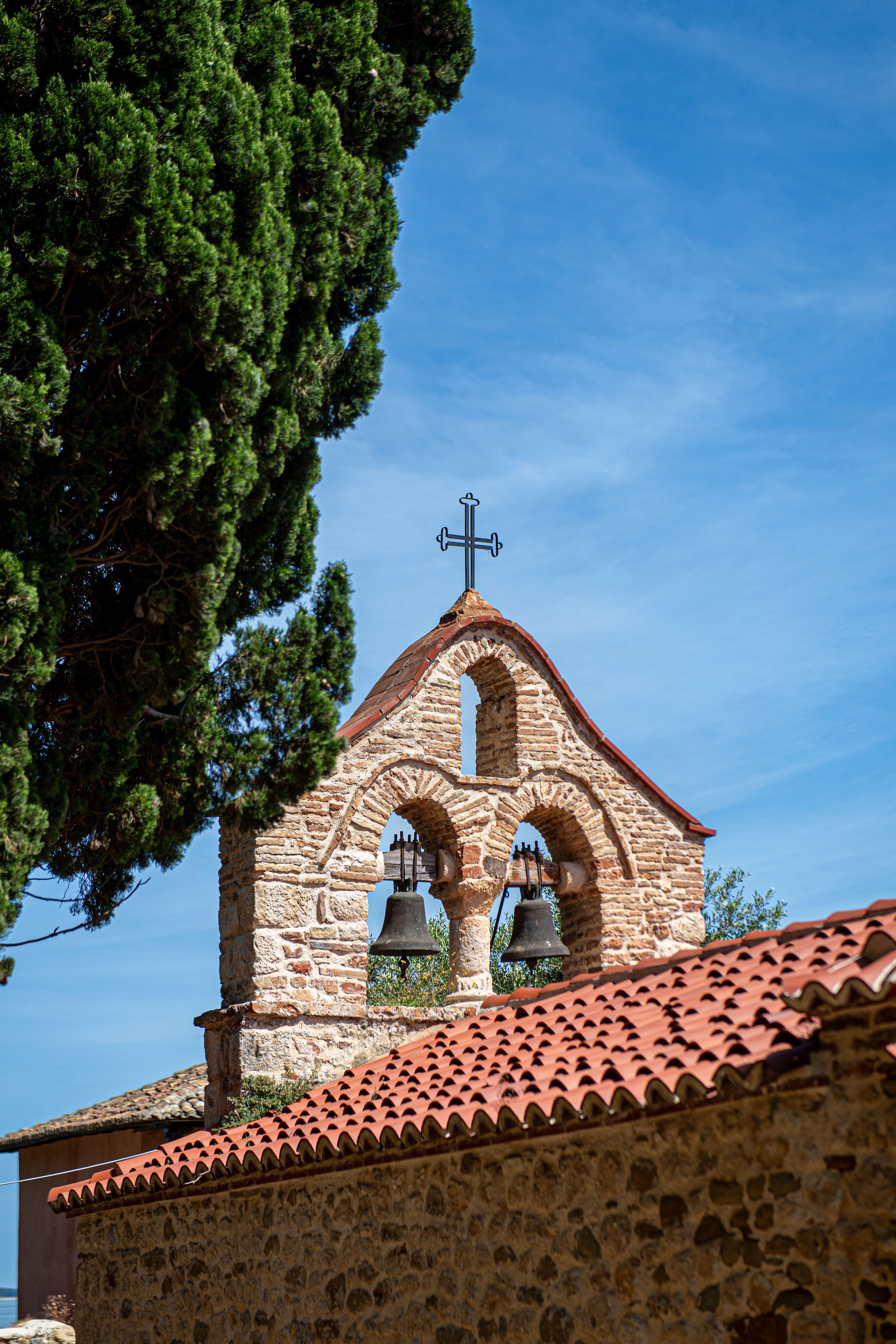 A rustic stone church bell tower with two large bells, a cross on top, and a tiled roof, set against a blue sky, with a large green tree nearby.