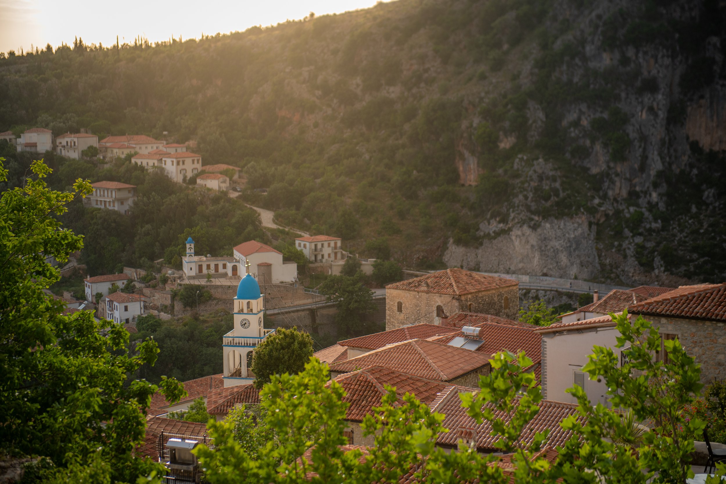 A scenic view of a small village with red-tiled roofs, a blue-domed church, and lush green trees, set against a hillside with rocky cliffs at sunset.