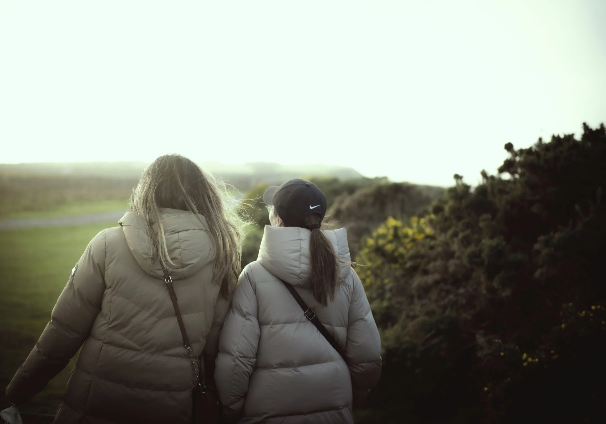 Two women with light-colored jackets, one with long hair and the other with a ponytail, walk outdoors during daytime near bushes and open fields.