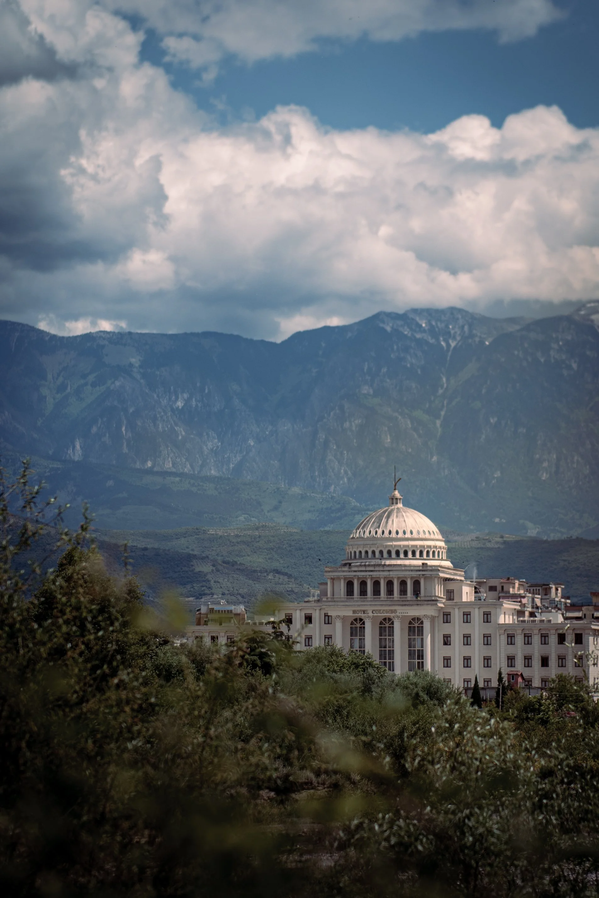 A white domed building labeled 'Hotel Colombia' in front of green trees, with mountains and a cloudy sky in the background.