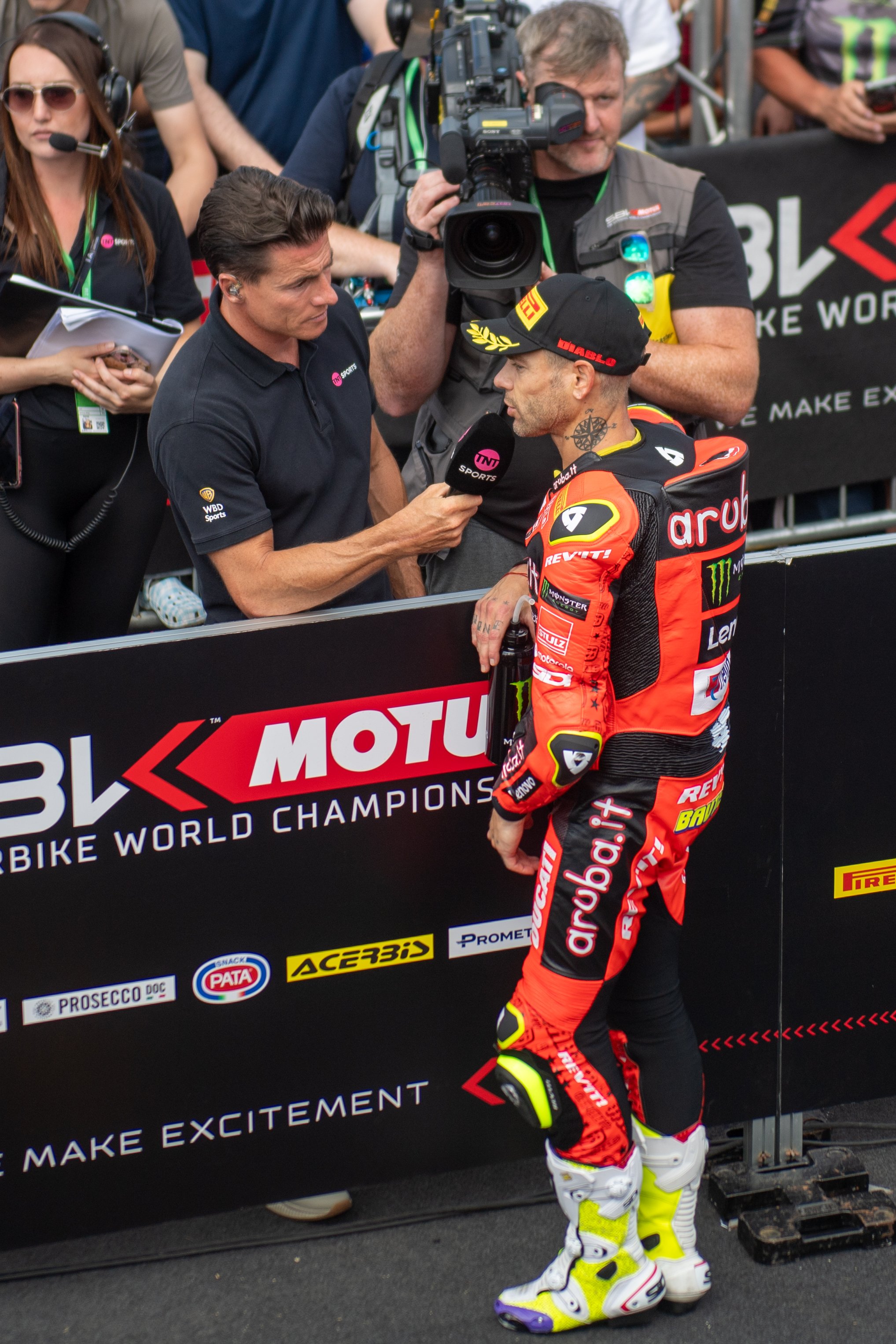 A motorcycle racer wearing a red and black racing suit and white racing boots is being interviewed by a man holding a microphone at a MotoGP event. The scene is at a paddock or trackside with other crew members and media personnel present.