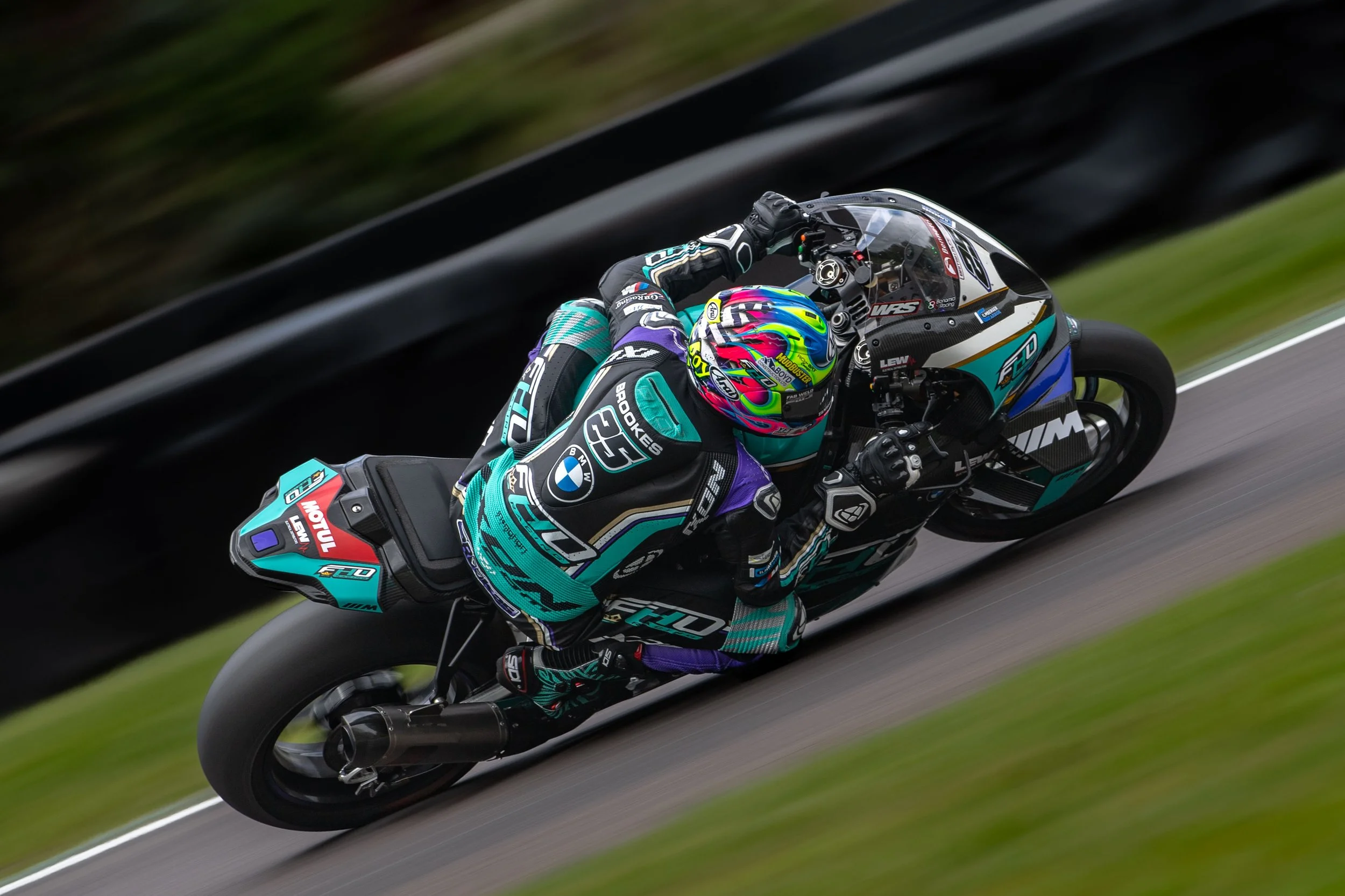 Motorcycle racer in a teal and black racing suit leaning into a turn on a racetrack, riding a sports motorcycle with a black and teal design and a colorful helmet.