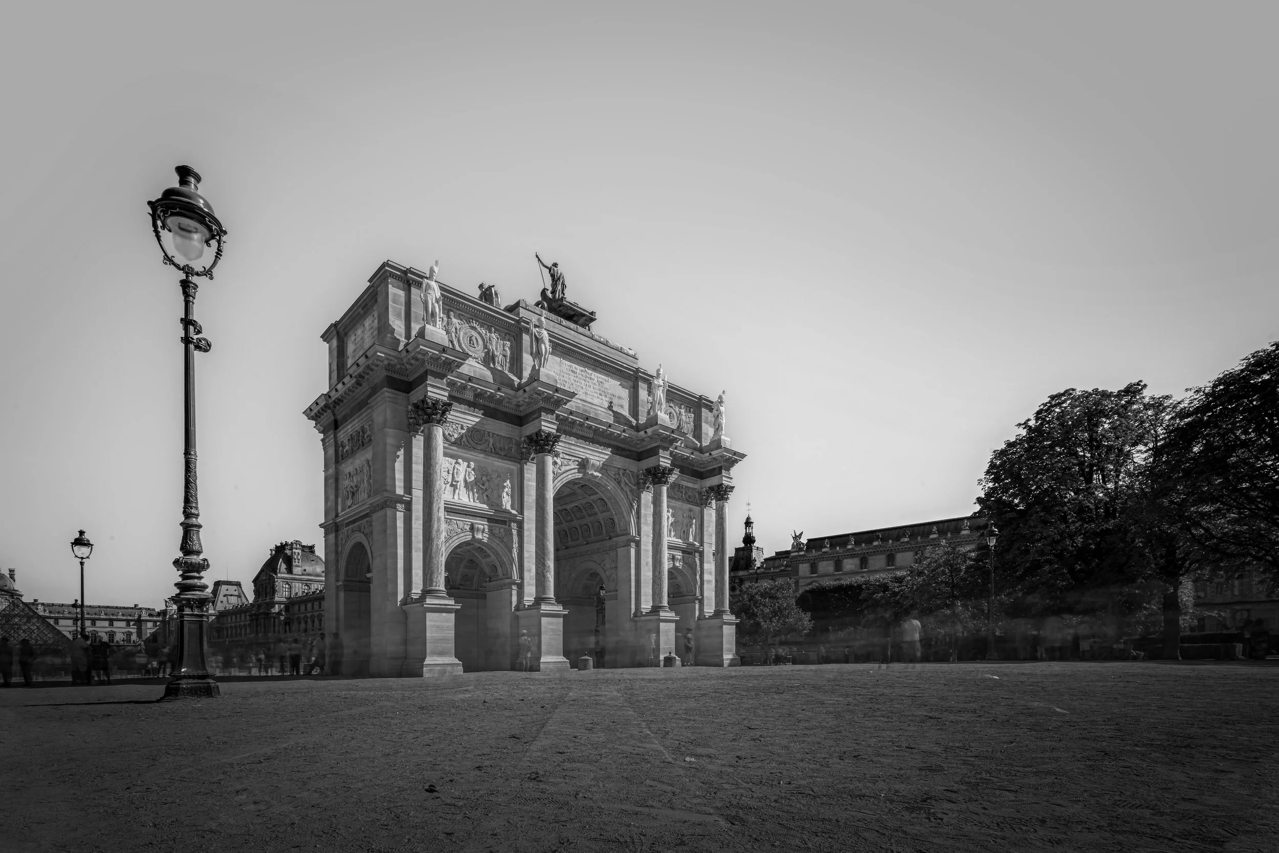Black and white photograph of Arc de Triomphe du Carrousel in Paris, France, with ornate details and sculptures. A streetlamp is visible in the foreground, along with trees and classical buildings in the background.