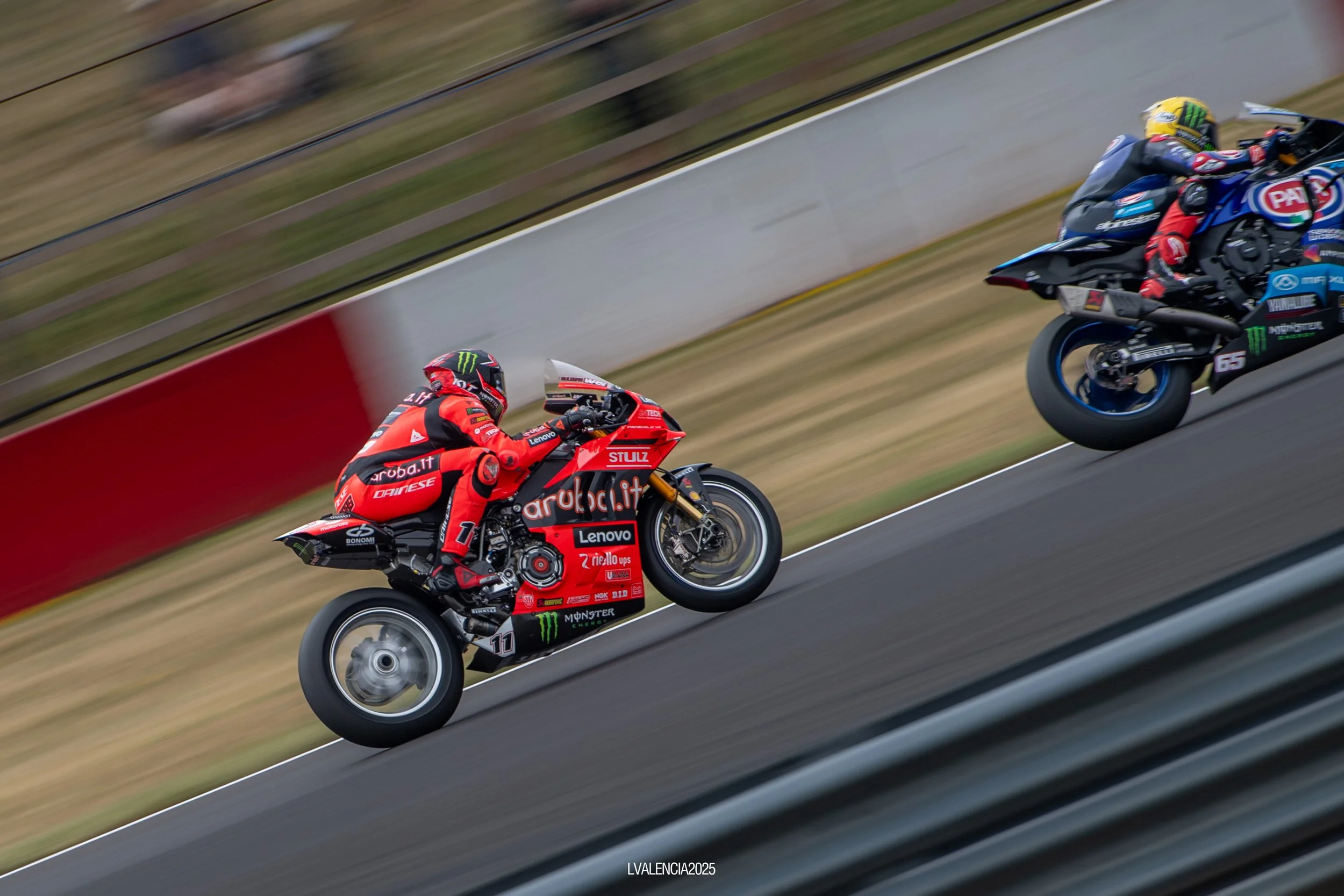 Two motorcycle racers in racing gear and helmets competing on a racetrack, with one rider on a red motorcycle and the other on a black and blue motorcycle, blurred background indicating high speed.