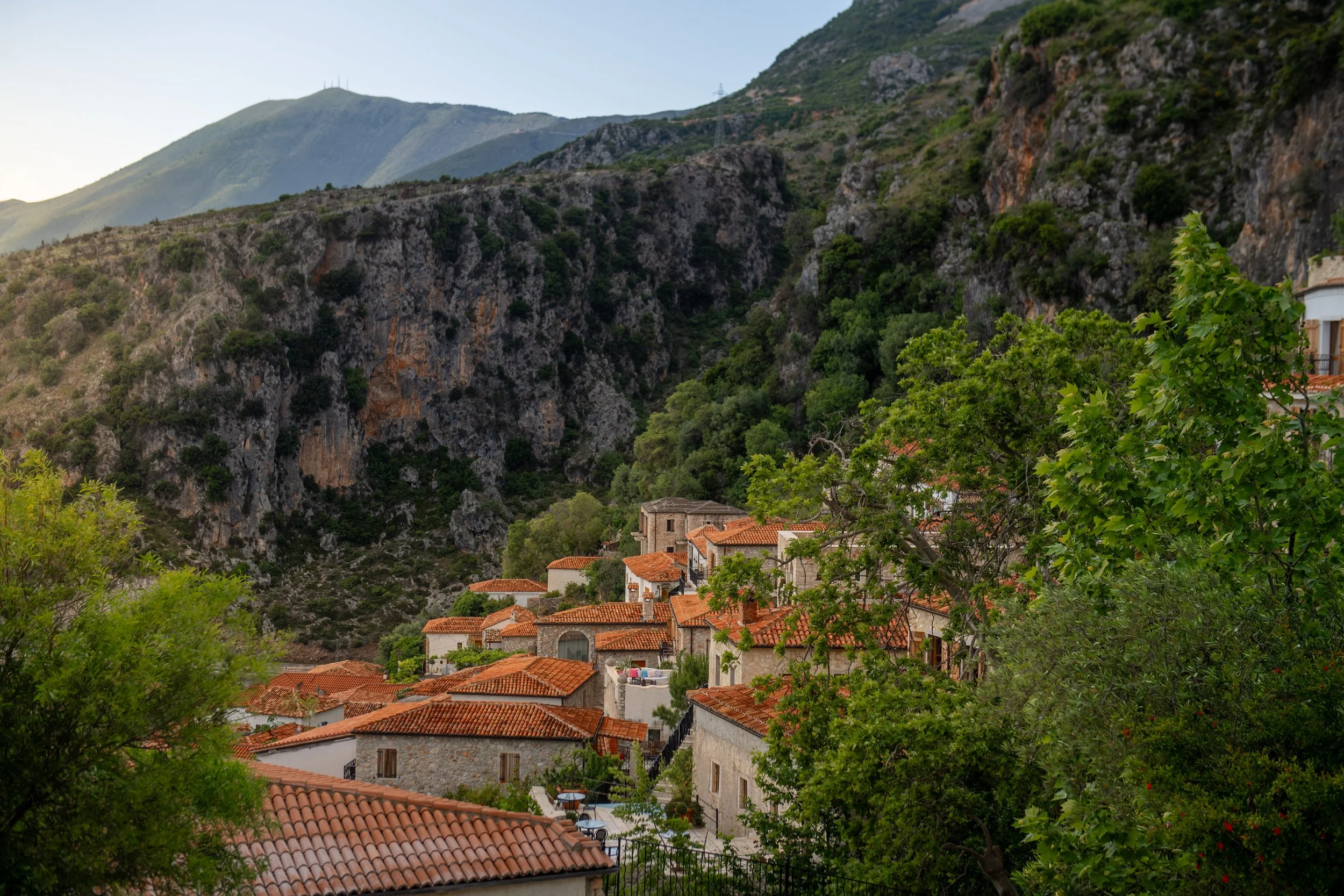 A small village nestled in a green mountain valley, with stone houses and red-tiled roofs surrounded by lush trees and rocky hillsides.
