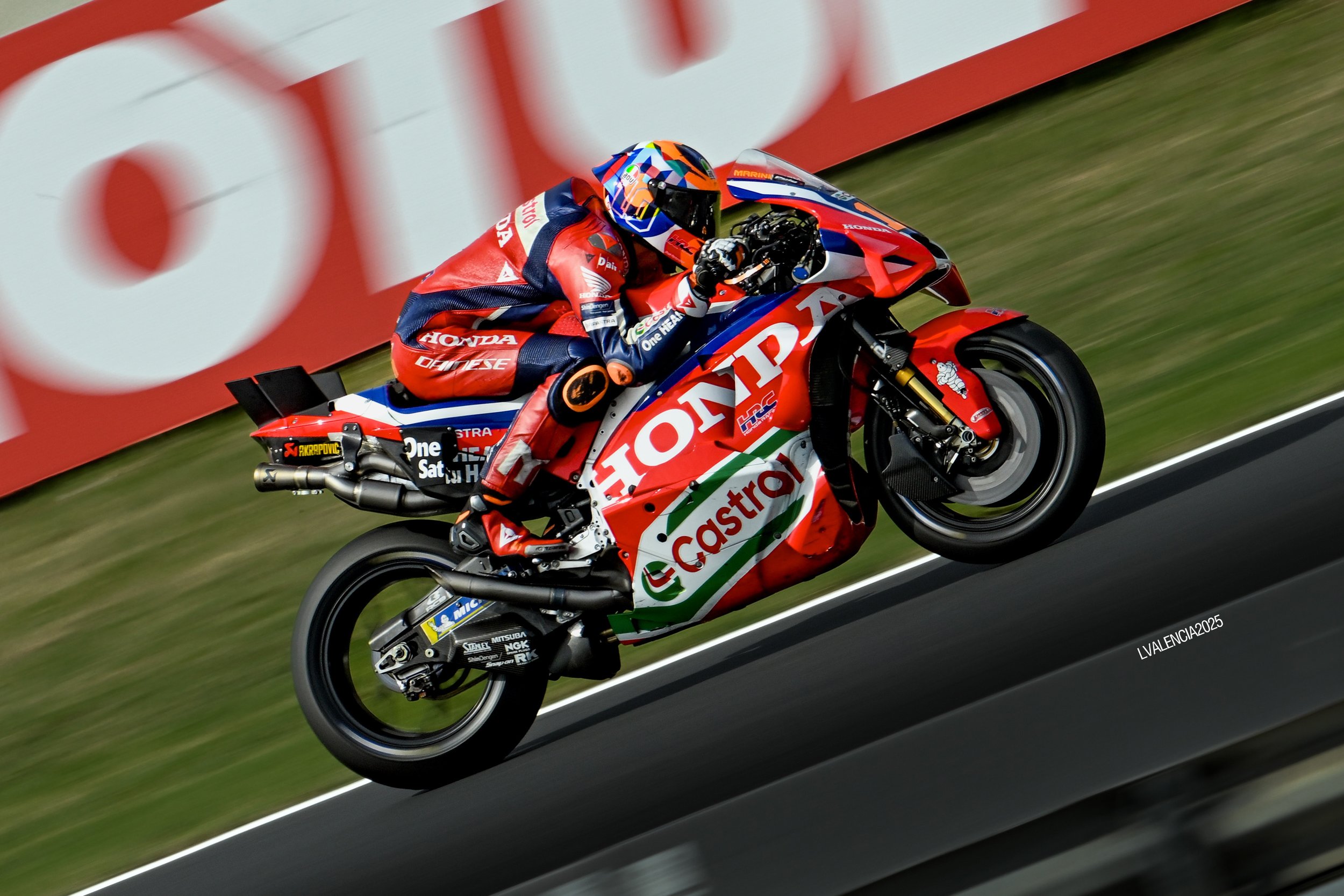 A professional motorcycle racer in a red, blue, and white racing suit riding a red Honda motorcycle with sponsor logos, including Castrol, on a race track.