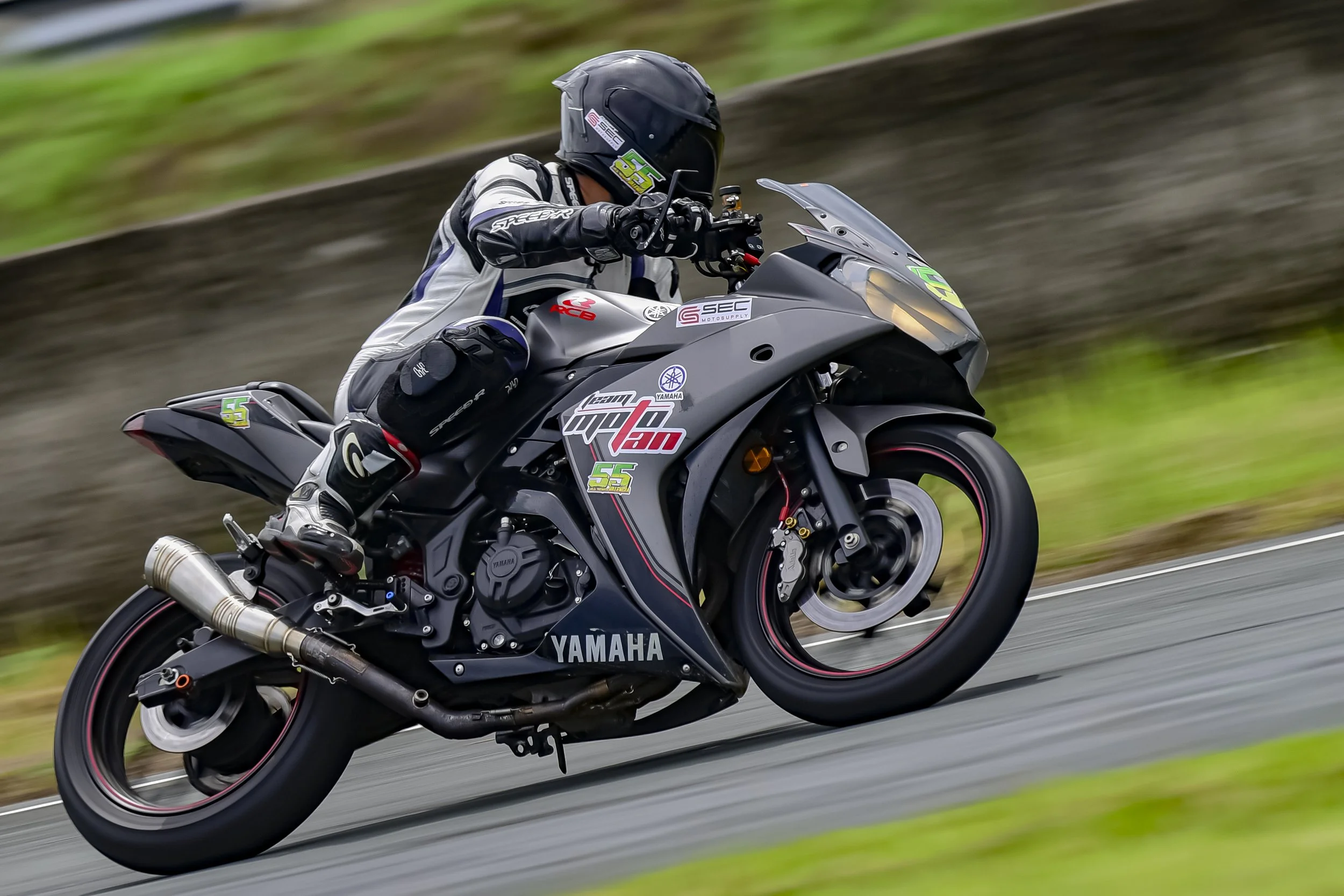 A motorcyclist racing on a Yamaha sport bike, leaning into a turn on a paved track with green grass and a gravel barrier in the background rider Lawrence Valencia