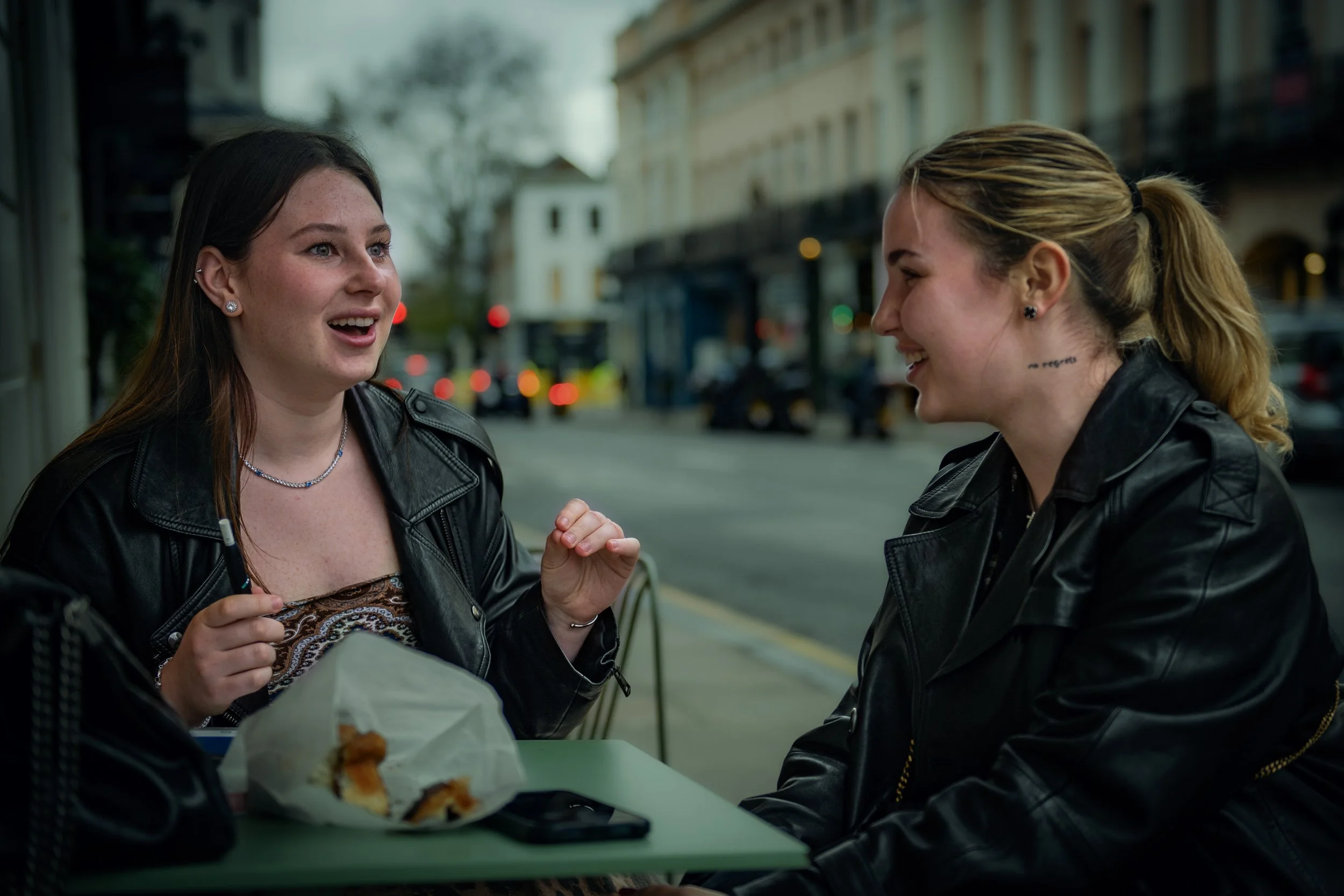 Two young women sitting at an outdoor café table on a city street, engaging in conversation and smiling. One is holding a pen and has a partially eaten pastry in front of her. Both are wearing black leather jackets.
