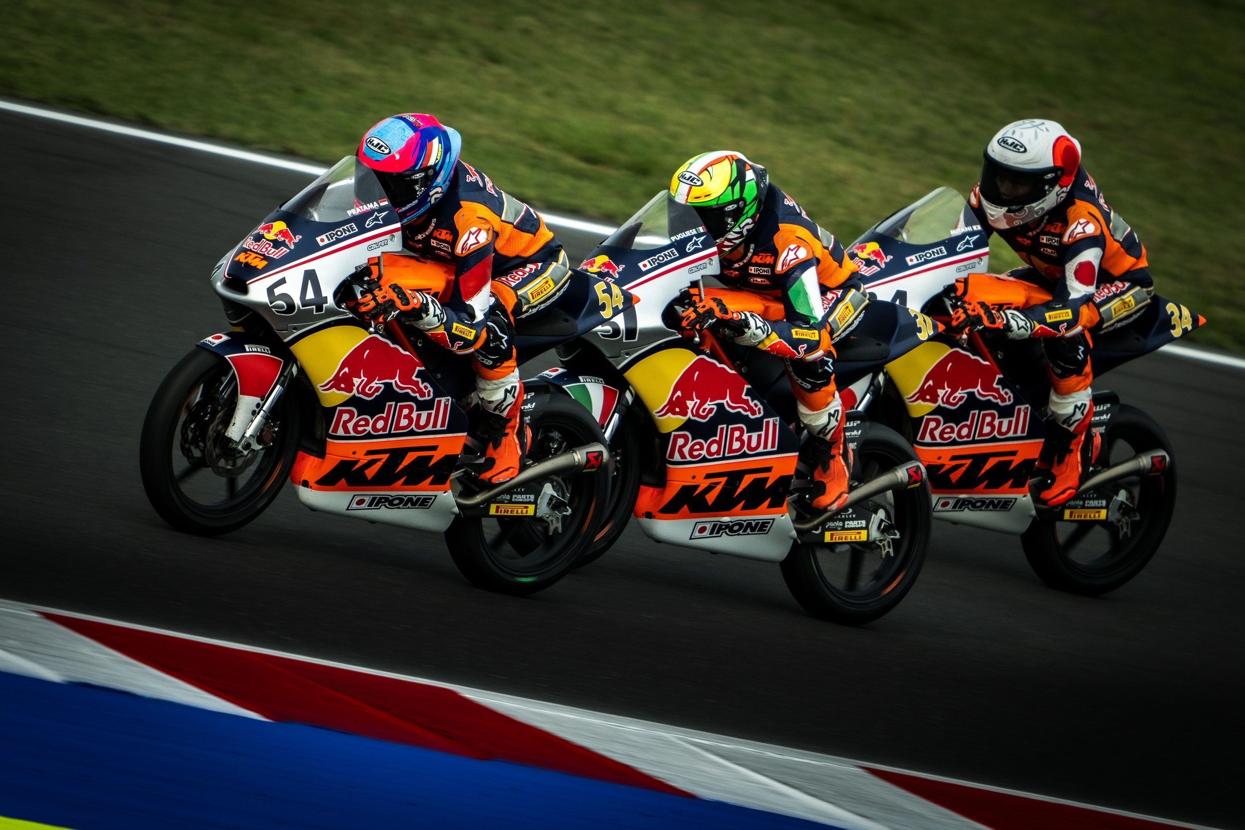 Three motorcycle racers in orange and black gear riding KTM bikes with Red Bull branding on a racetrack.