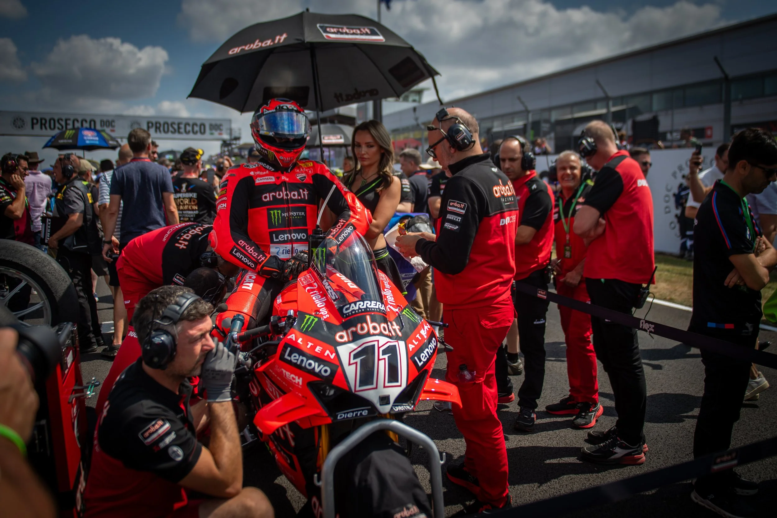 Race car driver in red racing suit sitting on a red motorcycle in the pit area surrounded by team members, photographers, and media personnel at the racetrack.