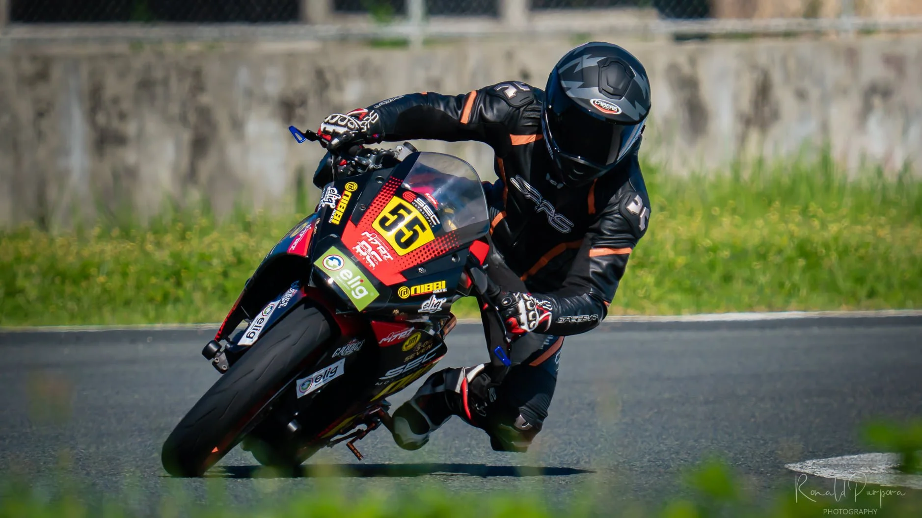 Motorcycle racer wearing black and orange gear leaning into a turn on a racetrack, with the number 55 on the front of the bike. rider name Lawrence Valencia