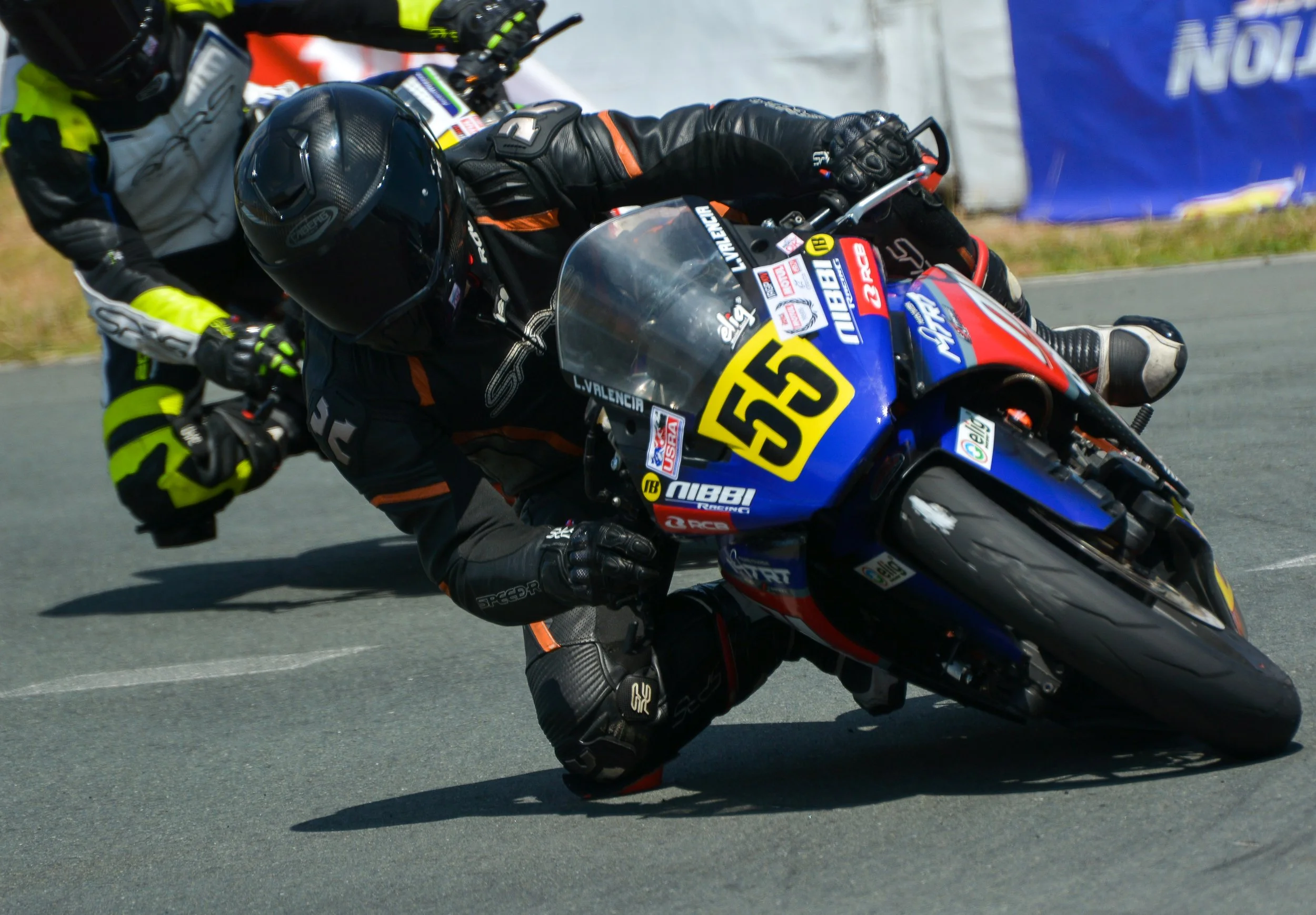 Motorcycle racer leaning into a sharp turn on the racetrack wearing black and orange gear and a black helmet, with the rider's number 53 visible on the front of the bike. rider name Lawrence Valencia