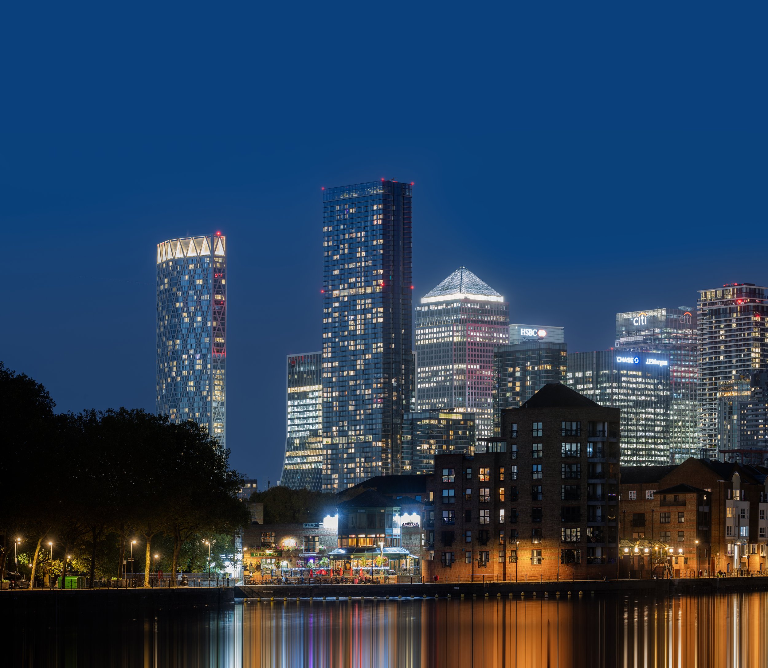 Nighttime city skyline with illuminated skyscrapers reflecting on water, with trees and a park in the foreground.