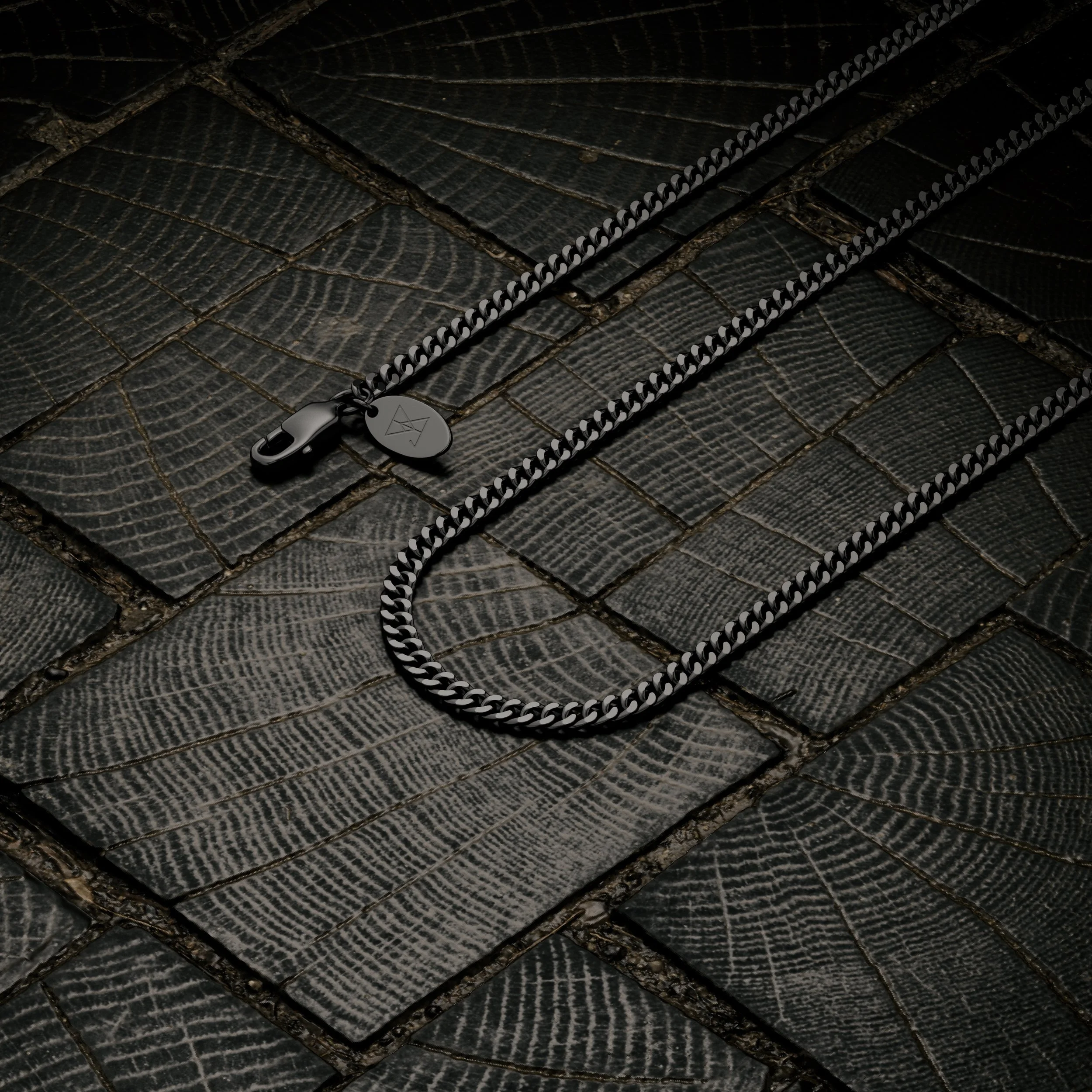 Close-up of a black and silver chain with a clasp and a small tag, lying on textured black tiles resembling tree rings.